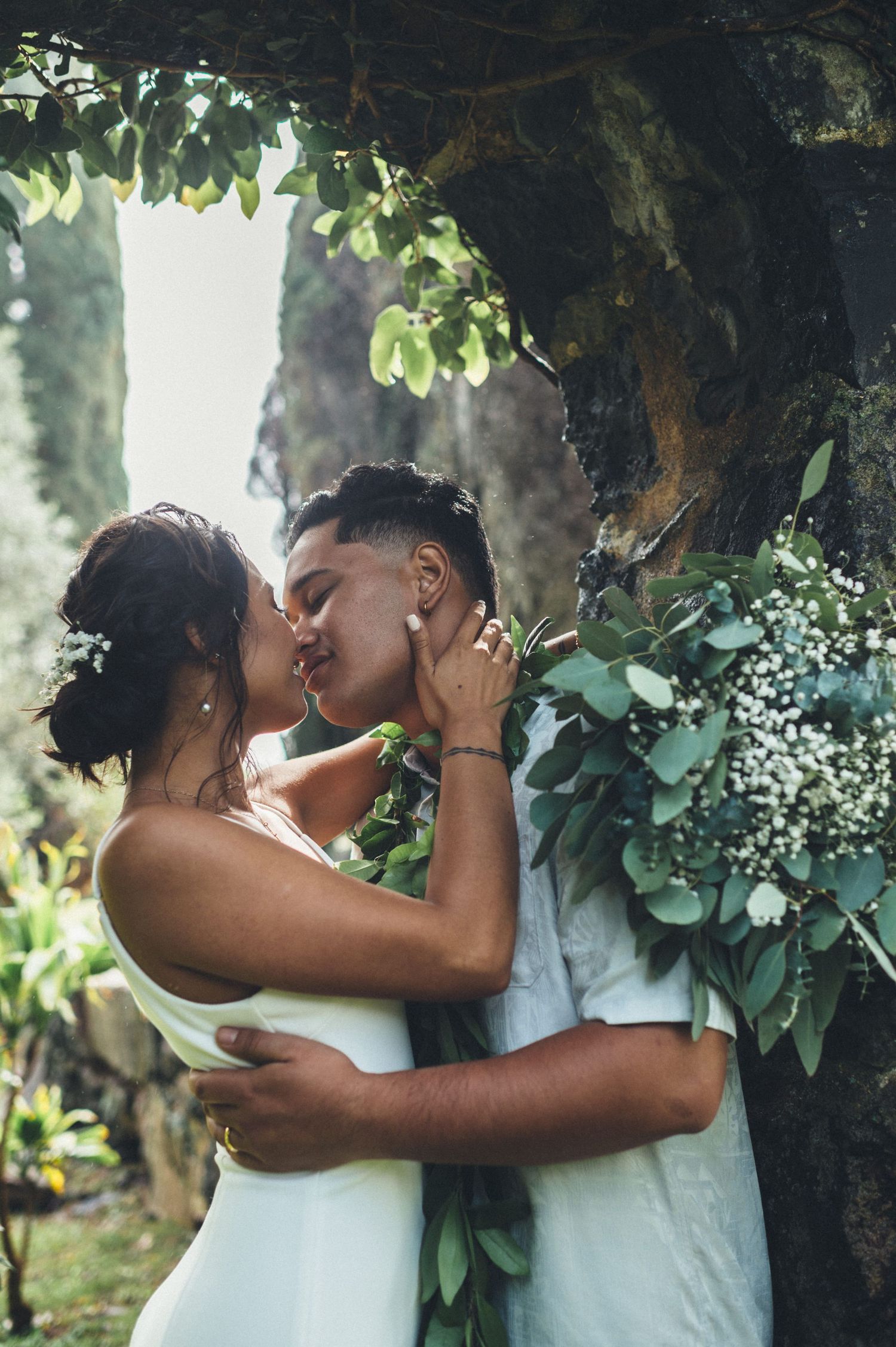 Couple sharing a romantic kiss under a tree, bride in white dress holding a eucalyptus bouquet with baby's breath.