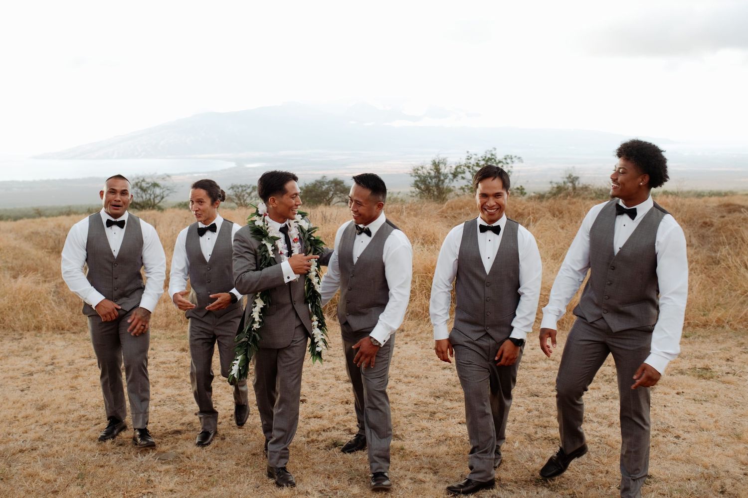 Groom and groomsmen in gray vests and bow ties walking outdoors at a scenic Hawaiian wedding venue.