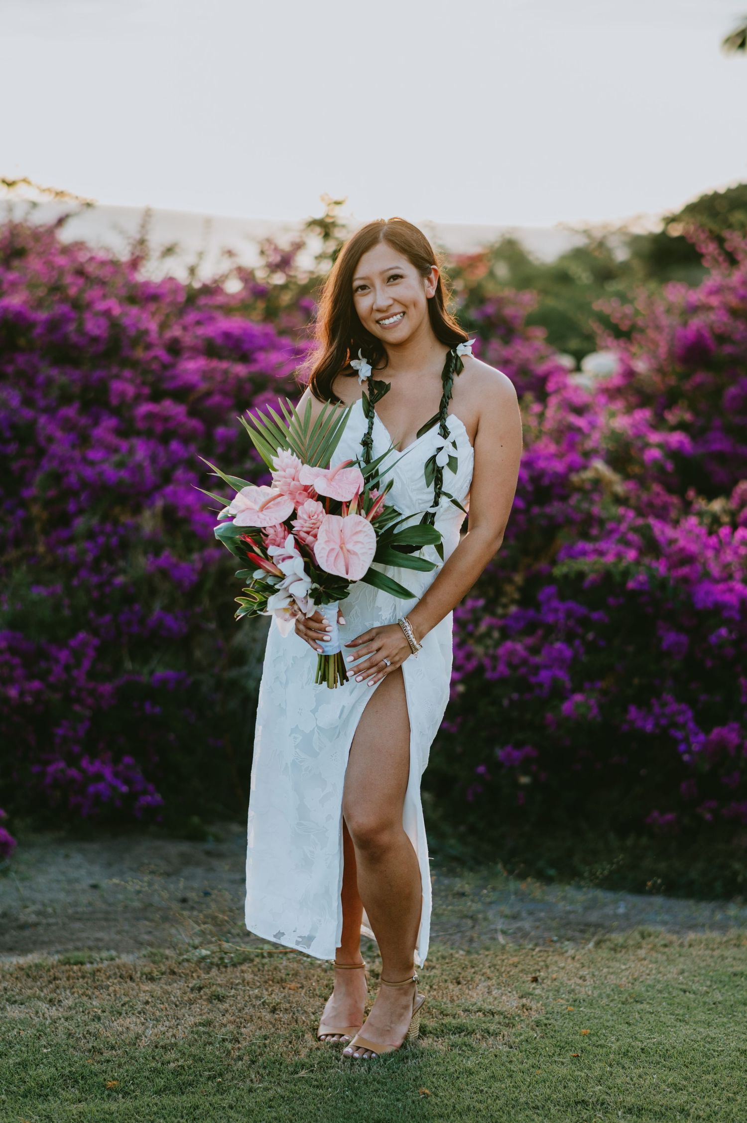Smiling bride in white slit dress holding tropical pink bouquet, standing before vibrant purple bougainvillea at sunset.
