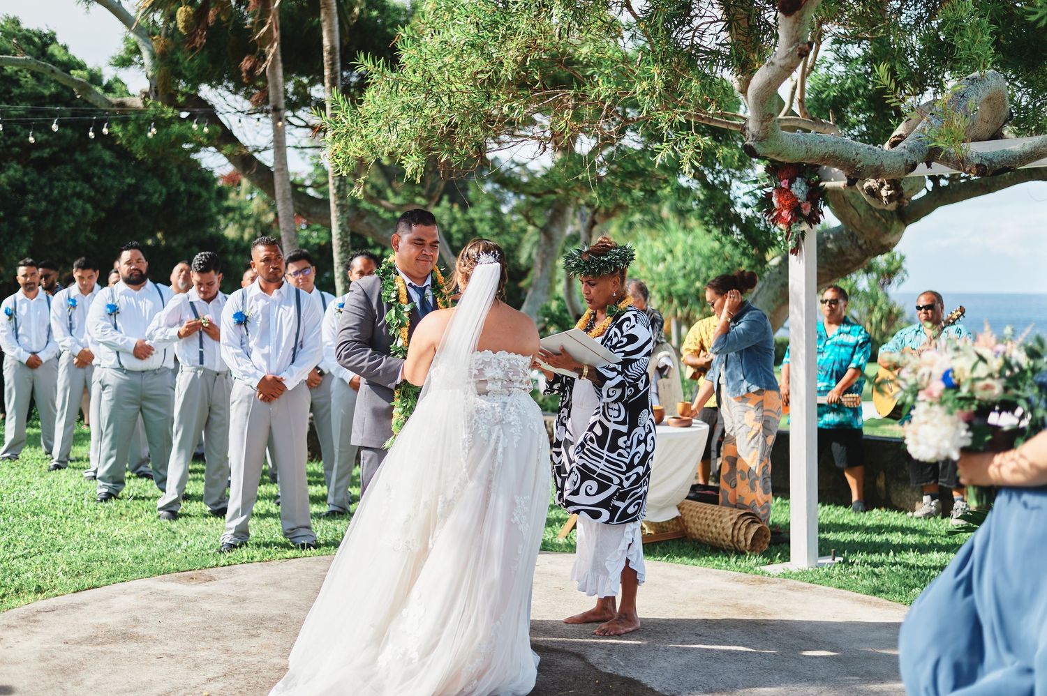 Outdoor wedding ceremony takes place under large trees with ocean view.