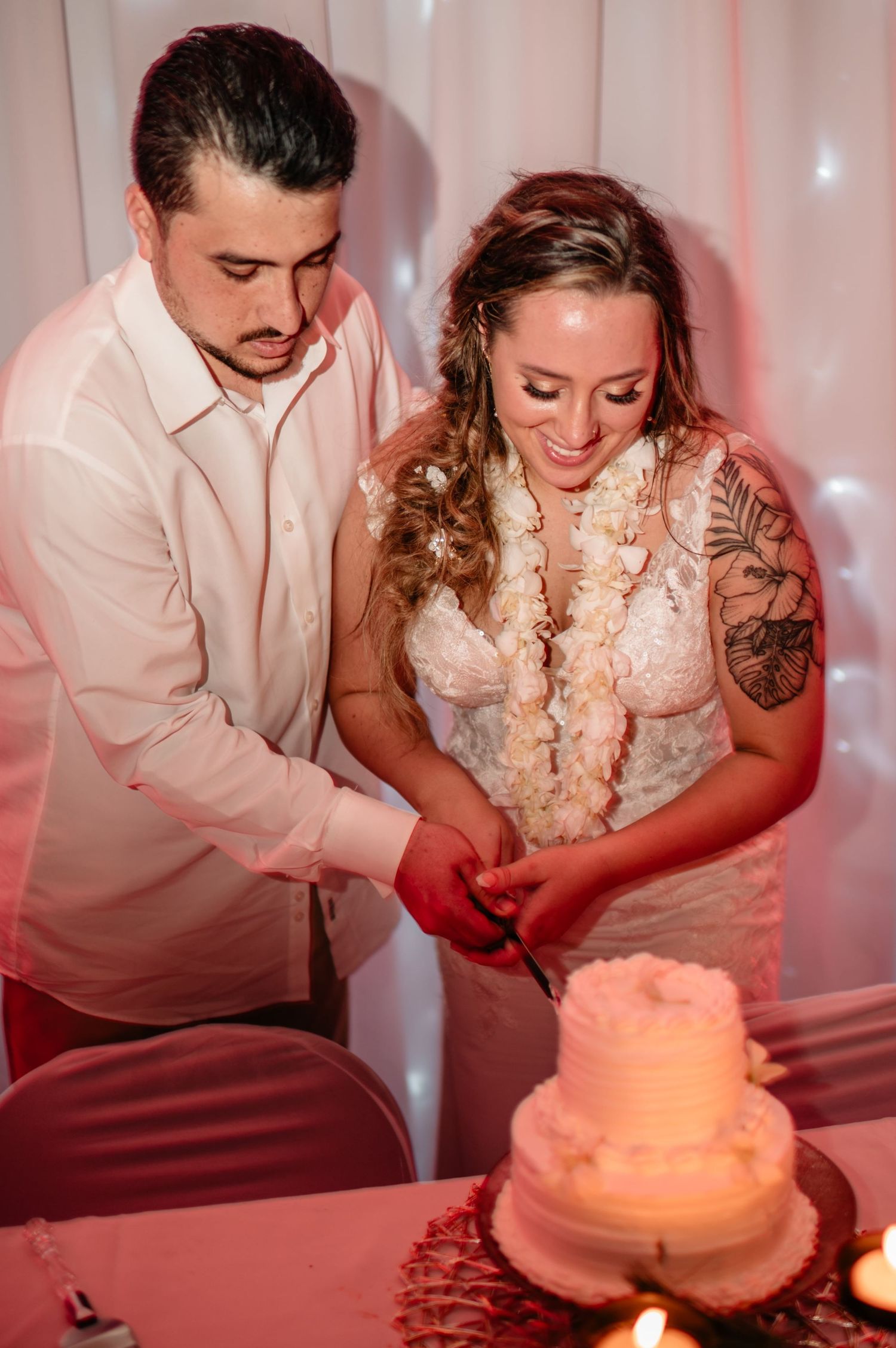 Couple cutting their wedding cake together at reception with pink lighting and white backdrop.
