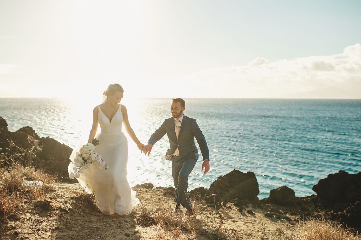 Bride and groom holding hands walking along rocky ocean cliffside at sunset in wedding attire.