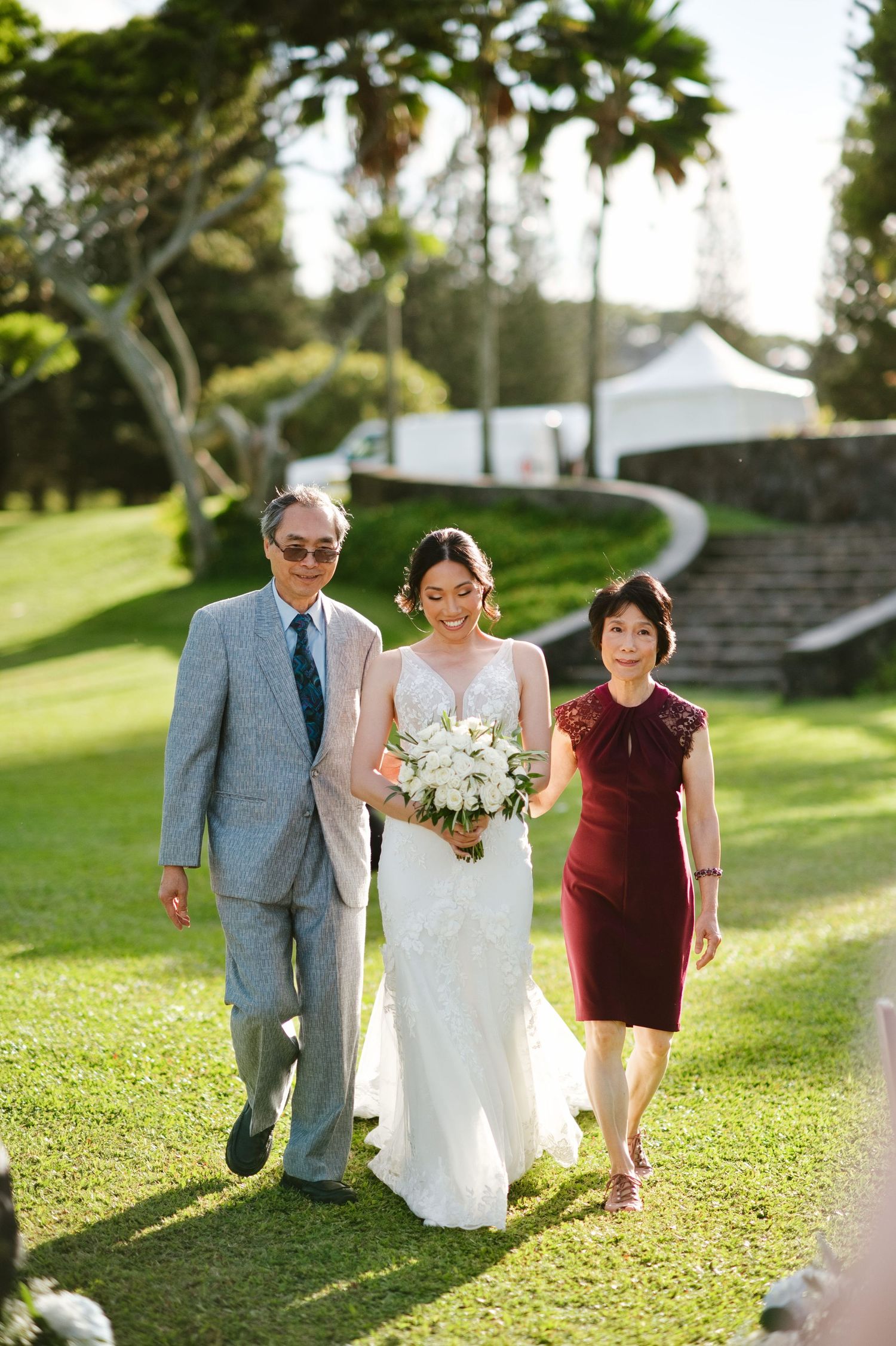 Bride in white gown walks with parents on green lawn, holding white bouquet, at outdoor wedding ceremony.