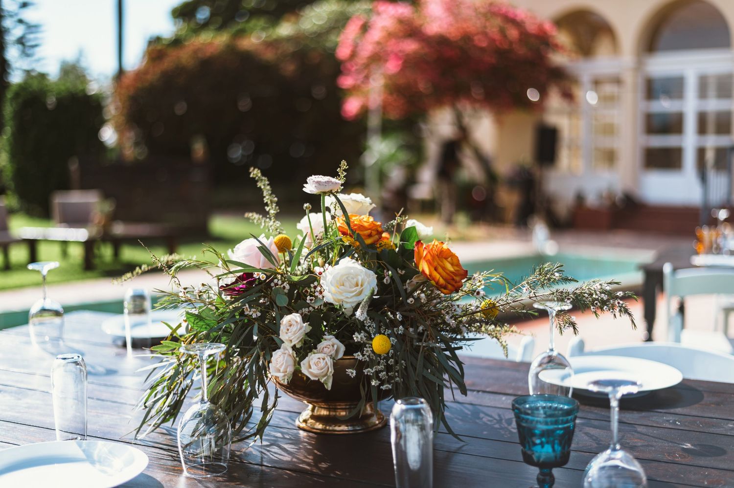 Elegant outdoor table setting with a lush floral centerpiece of orange and white blooms near a pool.