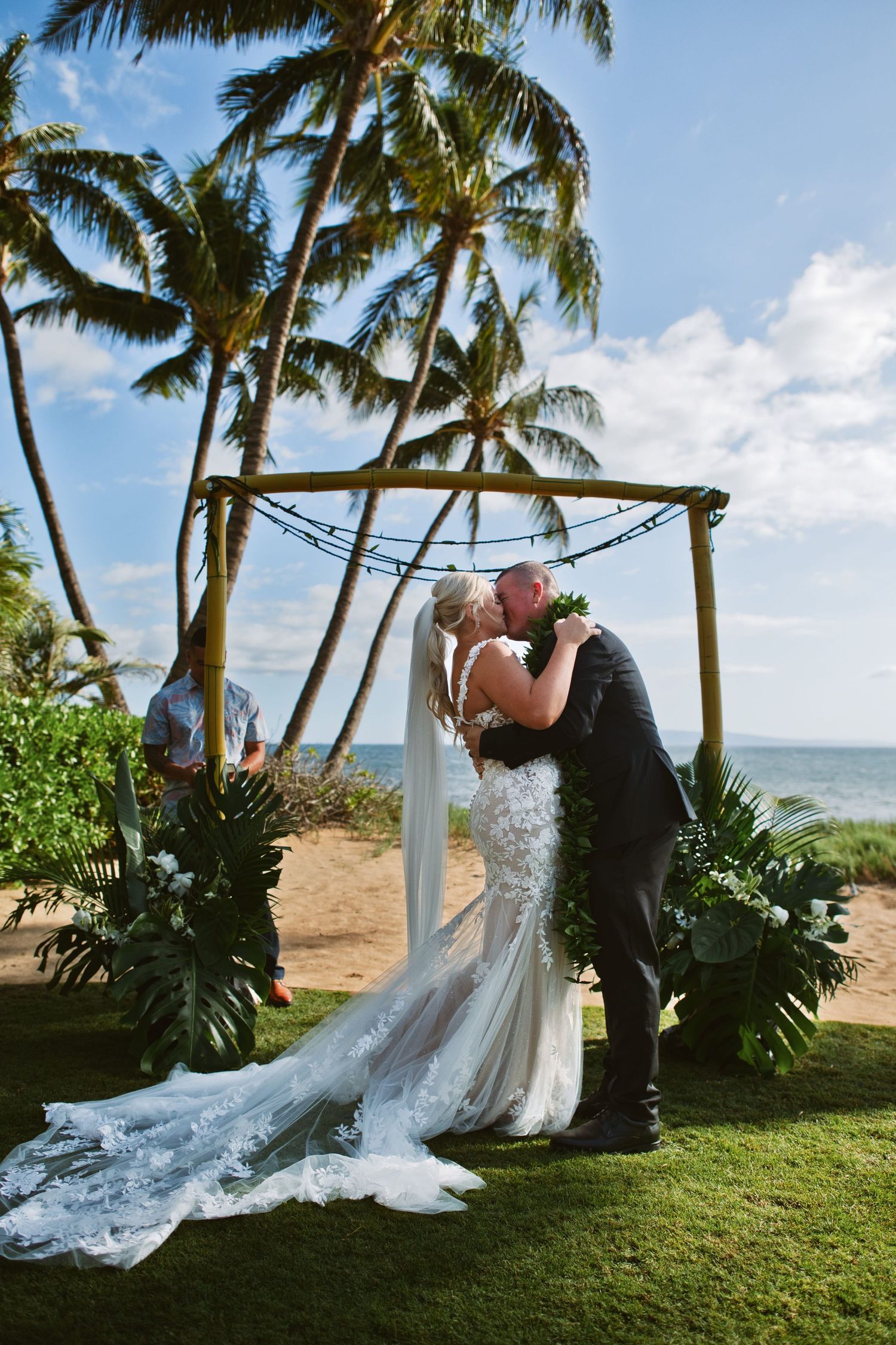 Bride and groom share first kiss at outdoor beach wedding ceremony under bamboo arch with tropical palm trees in Hawaii.