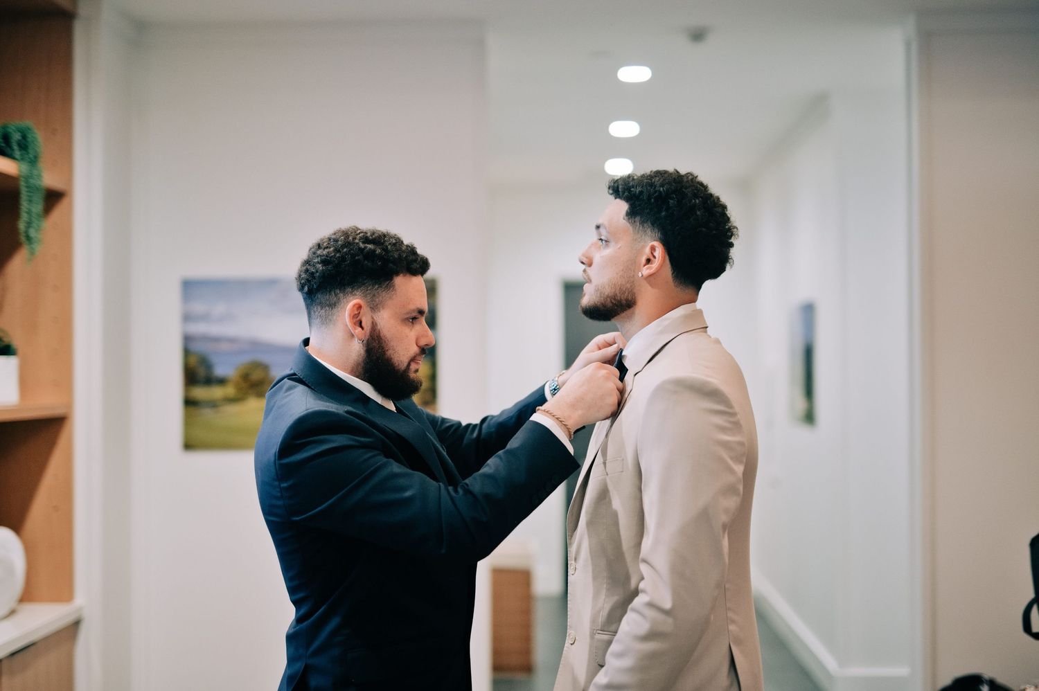 Two well-dressed men in suits adjusting a tie indoors, preparing for a formal event or wedding.