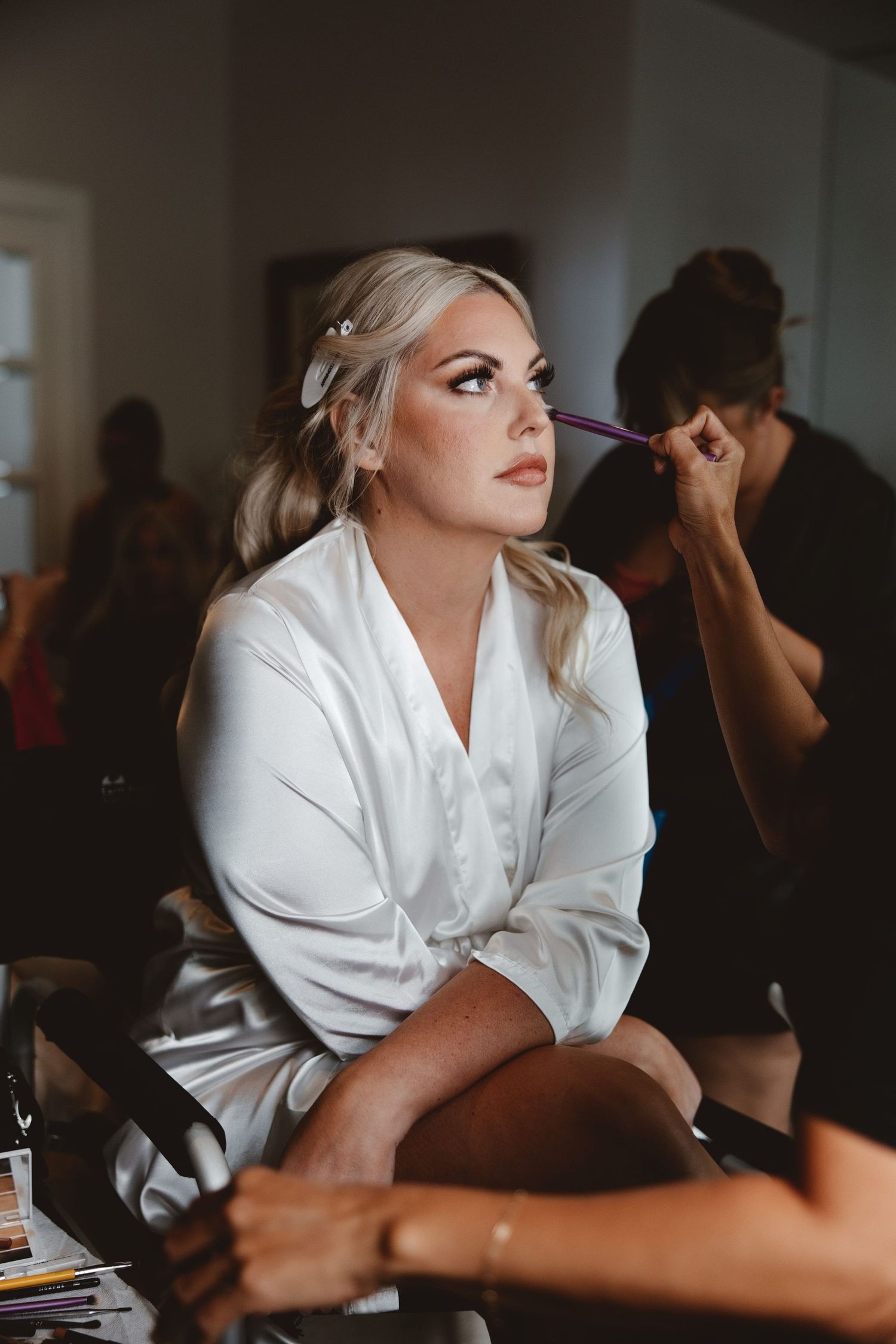 Bride in white satin robe getting makeup applied by artist during wedding morning preparations.