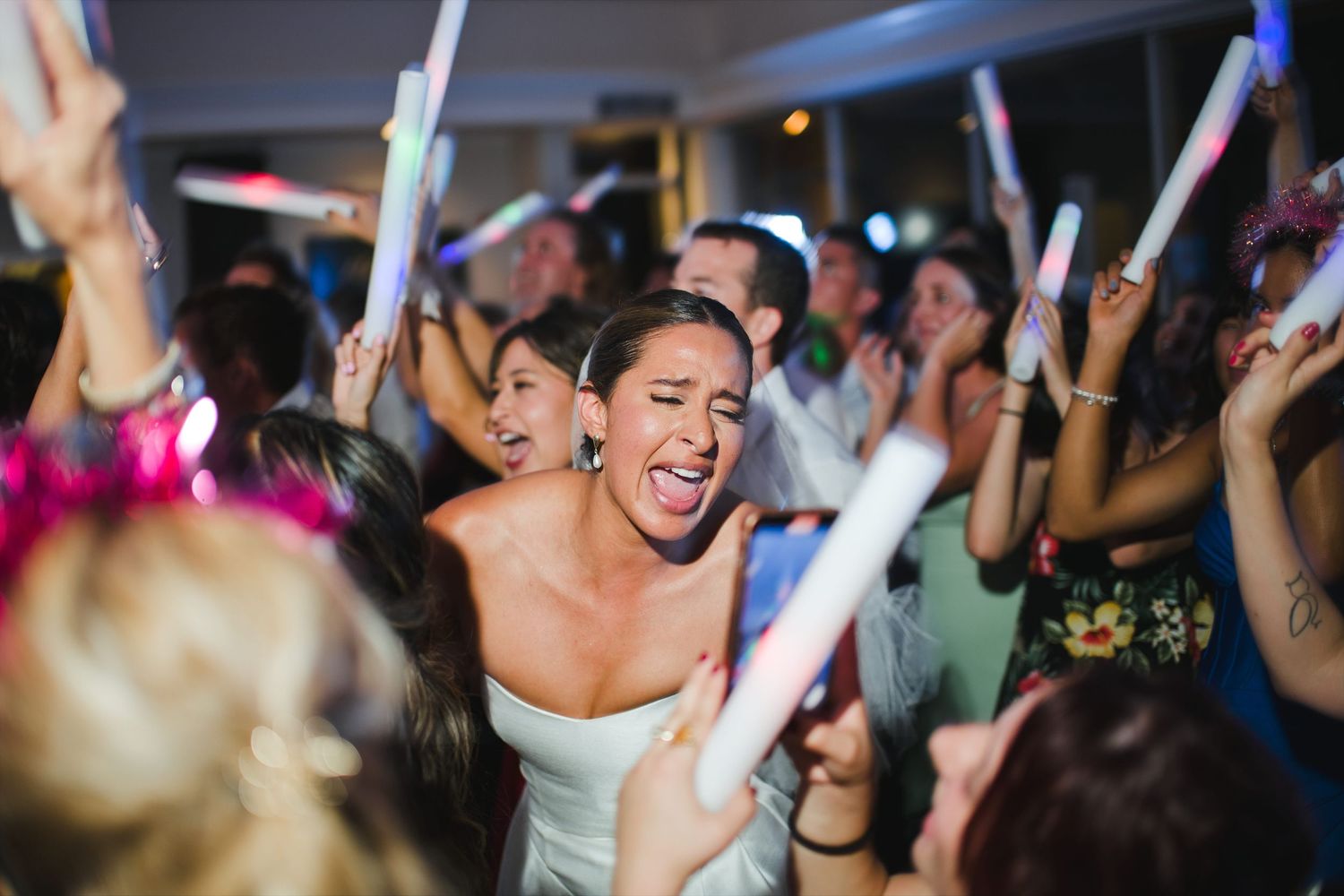 Bride dancing joyfully at wedding reception surrounded by guests holding glowing foam sticks.