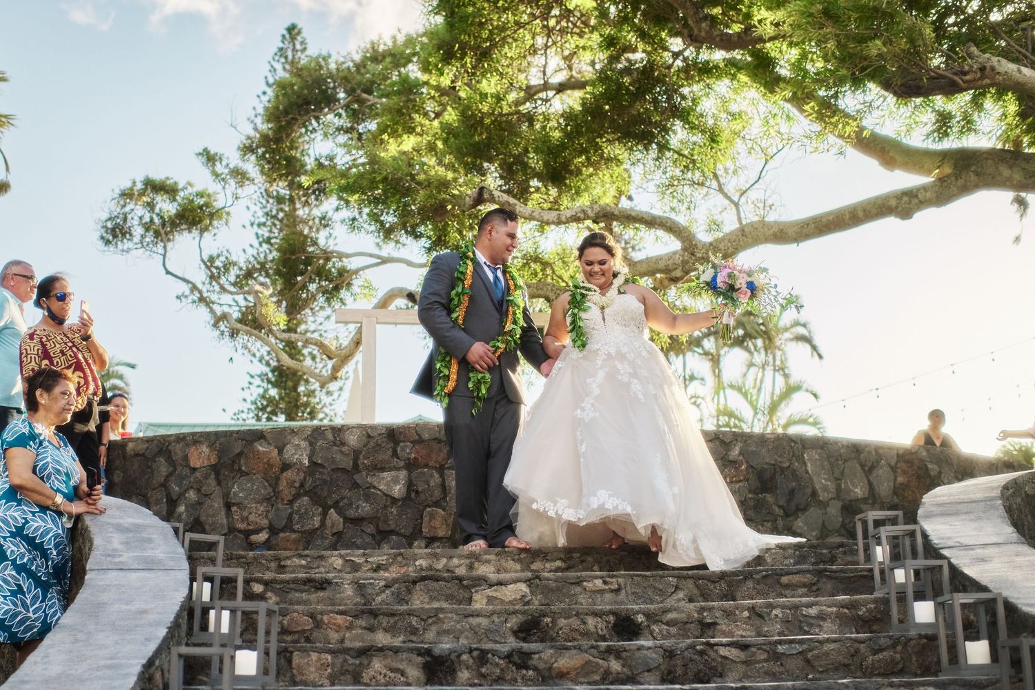 A wedding couple descends stone steps at an outdoor venue under a large tree.