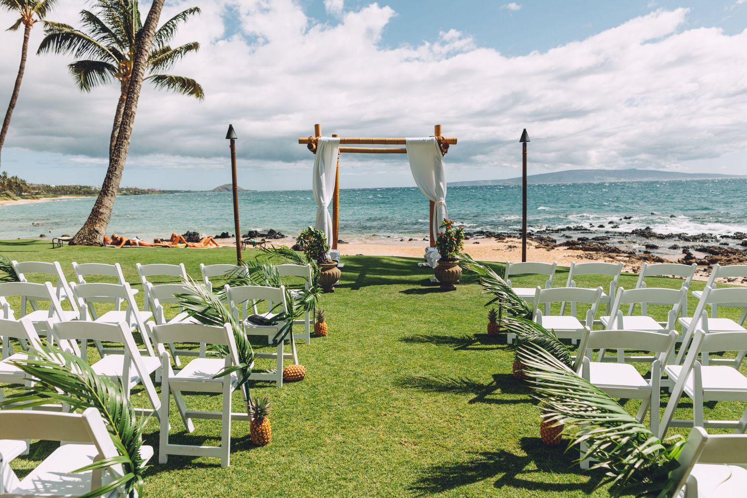Tropical beach wedding ceremony setup with white chairs and fabric arch overlooking the ocean.
