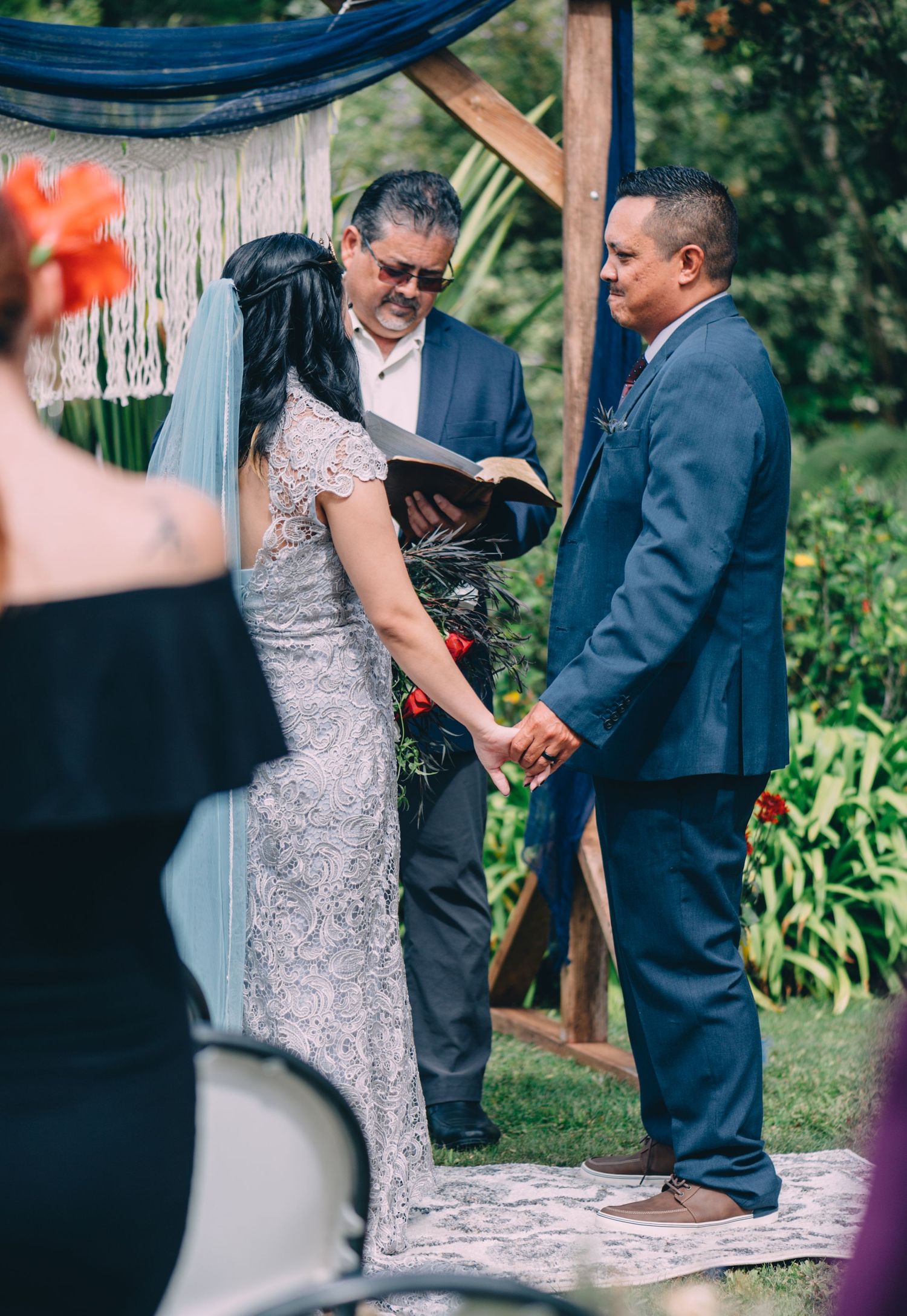 Wedding ceremony under wooden arch with guests seated in garden setting.
