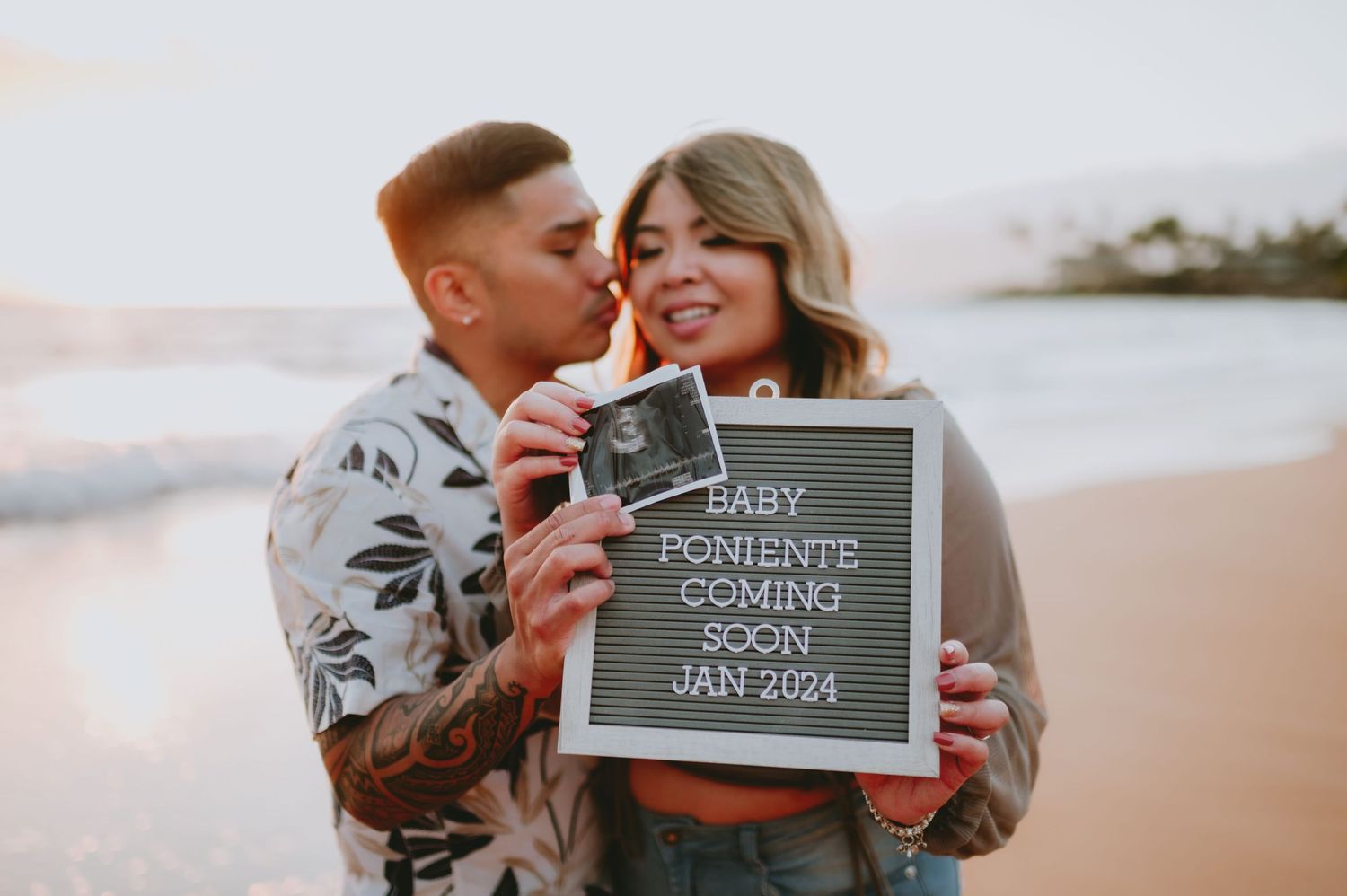 Couple on beach holding ultrasound and letter board sign announcing Baby Poniente coming soon Jan 2024.
