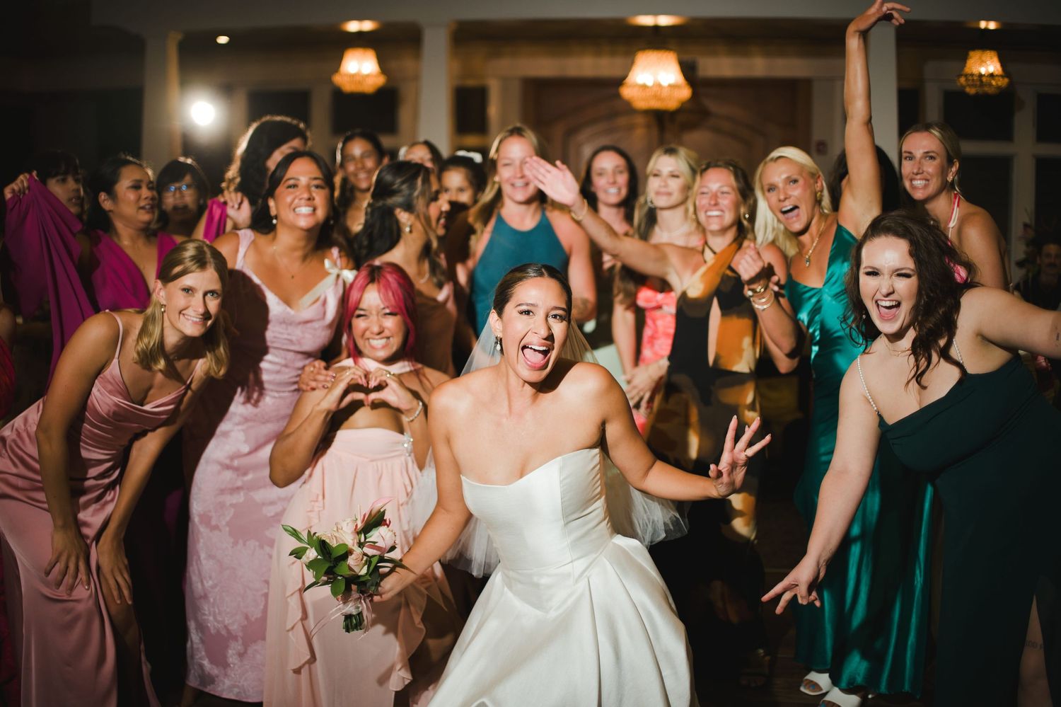 Bride laughing joyfully with excited bridesmaids in colorful dresses during wedding bouquet toss at elegant reception.
