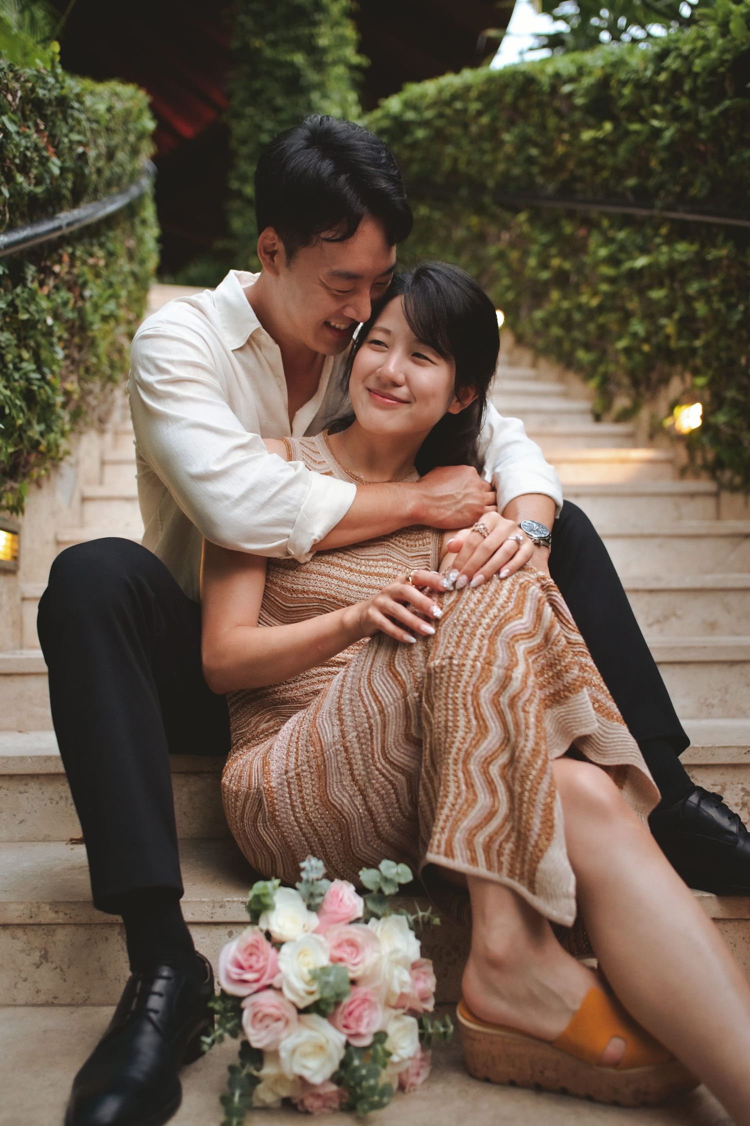 Couple sitting on outdoor steps, man embracing woman in knit dress, bouquet of pink roses nearby.