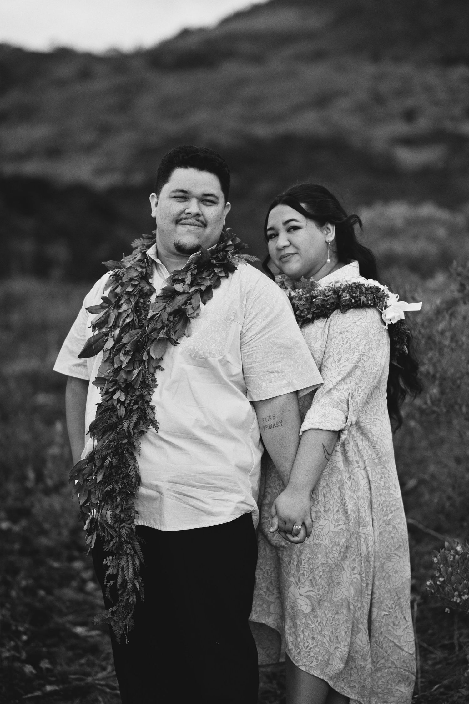 Black and white photo of a couple holding hands outdoors, wearing traditional Hawaiian leaf leis, smiling together.