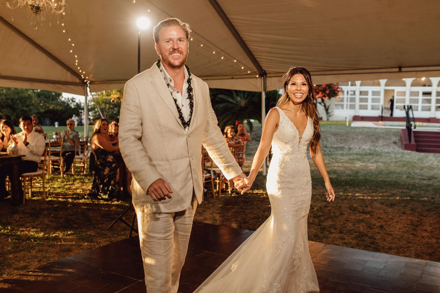 Bride and groom hold hands walking into outdoor wedding reception under white tent with string lights.
