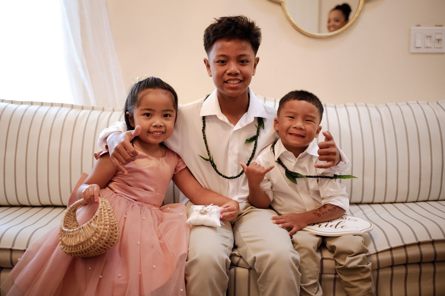 Three children in formal attire sit together on a sofa, smiling, with the older boy hugging the younger two.