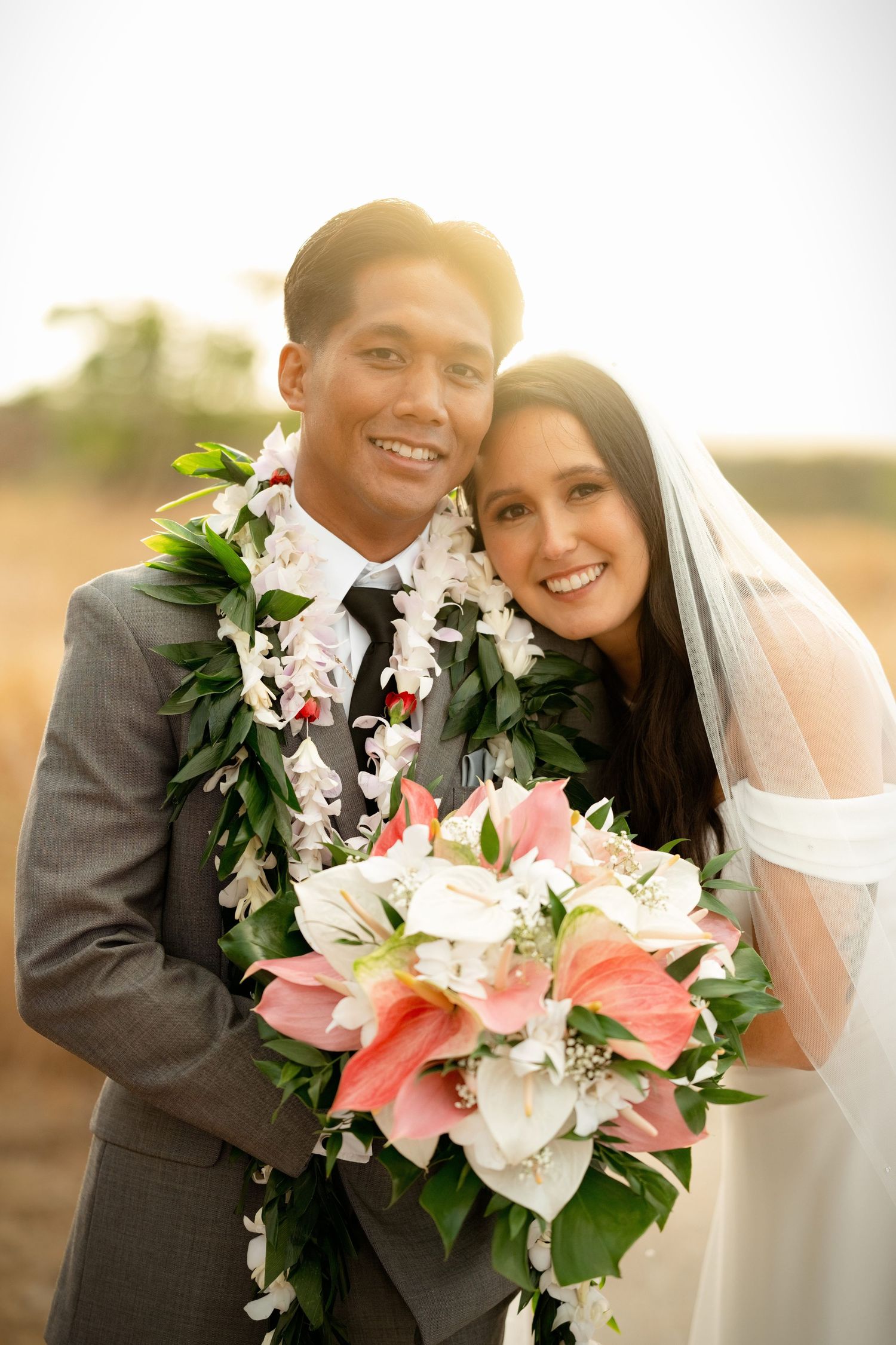 Smiling interracial newlywed couple posing with pink and white bouquet, groom wearing floral lei, bride in veil, outdoor sunset.