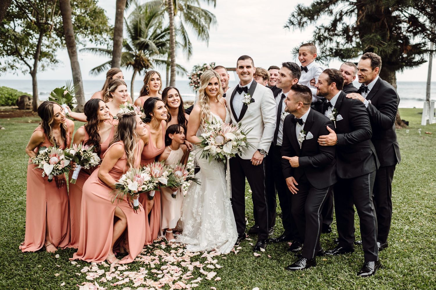 Wedding party poses outdoors, bridesmaids in coral gowns and groomsmen in black tuxedos with tropical palm trees background.