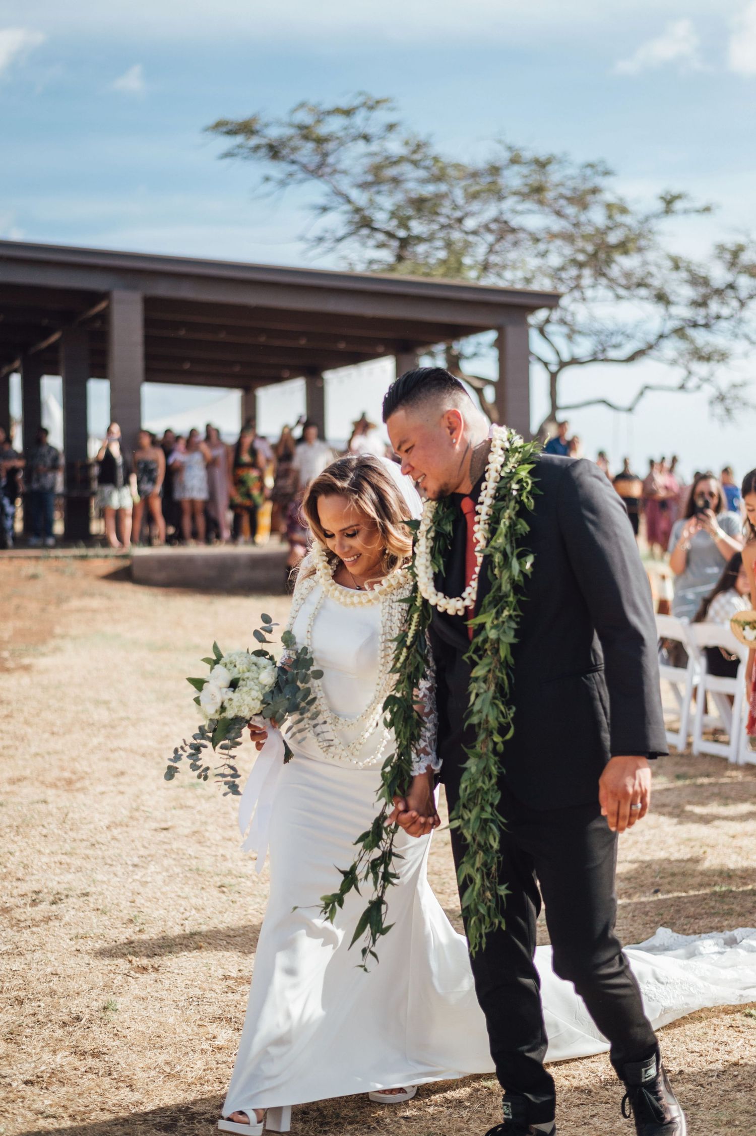 Newlywed couple walking down aisle after Hawaiian wedding ceremony, groom wearing green lei, bride in white gown holding bouquet.