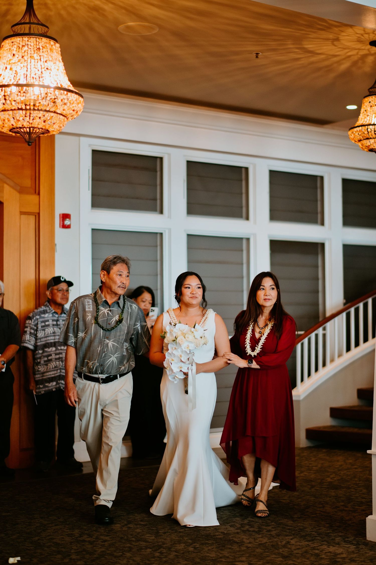 Bride in white gown walks with parents through elegant venue with chandeliers during wedding processional.