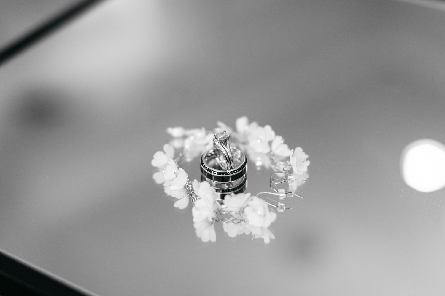 Black and white photo of wedding rings nestled in delicate white flowers on a reflective surface.