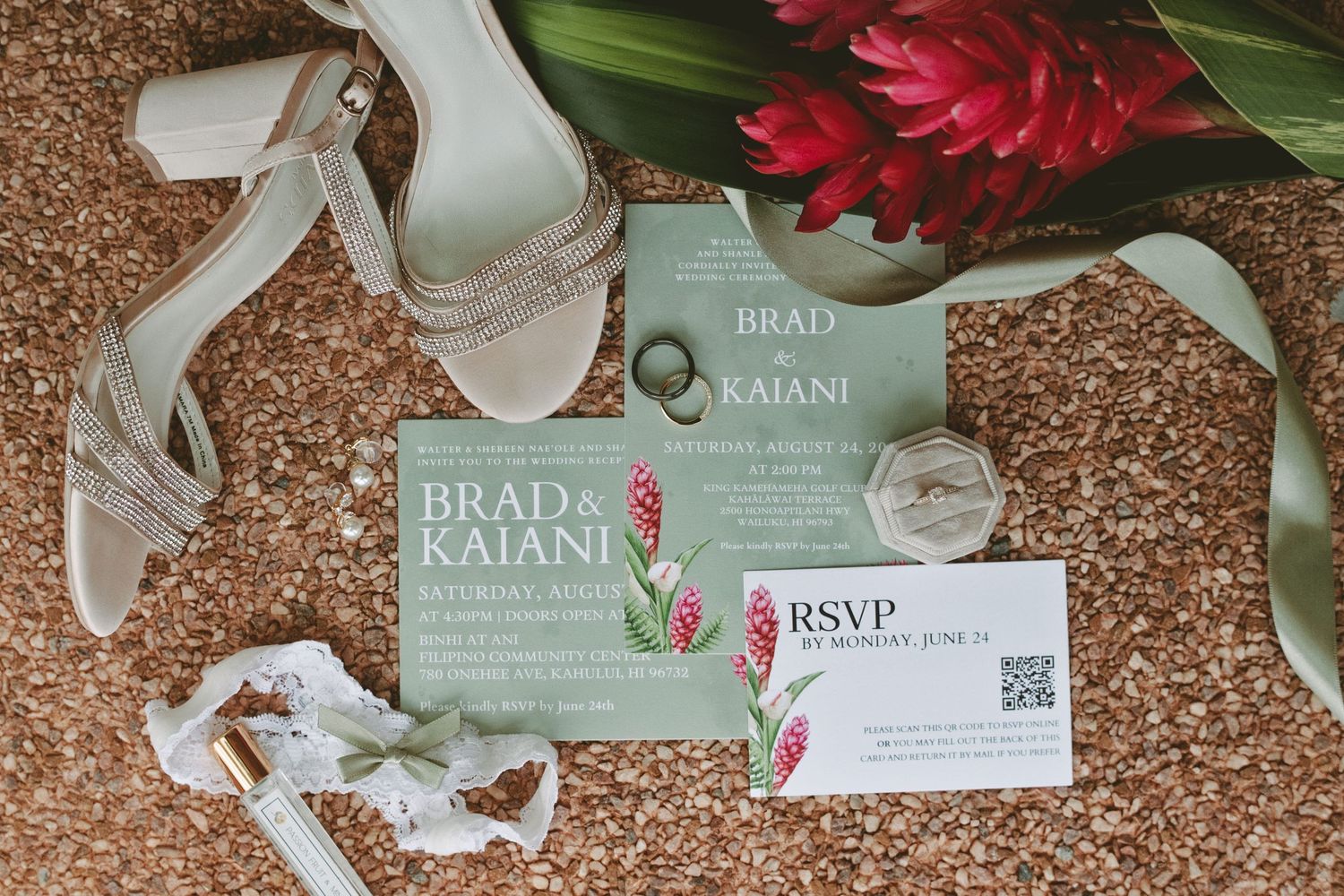 Wedding invitation suite for Brad and Kaiani displayed with bridal heels, rings, and tropical red ginger flowers on cork background.