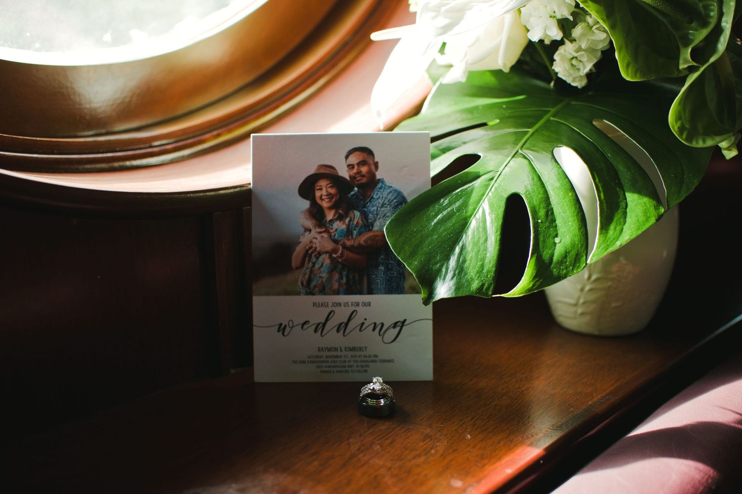 Wedding invitation card with couple photo, diamond ring, and monstera plant on wooden table.