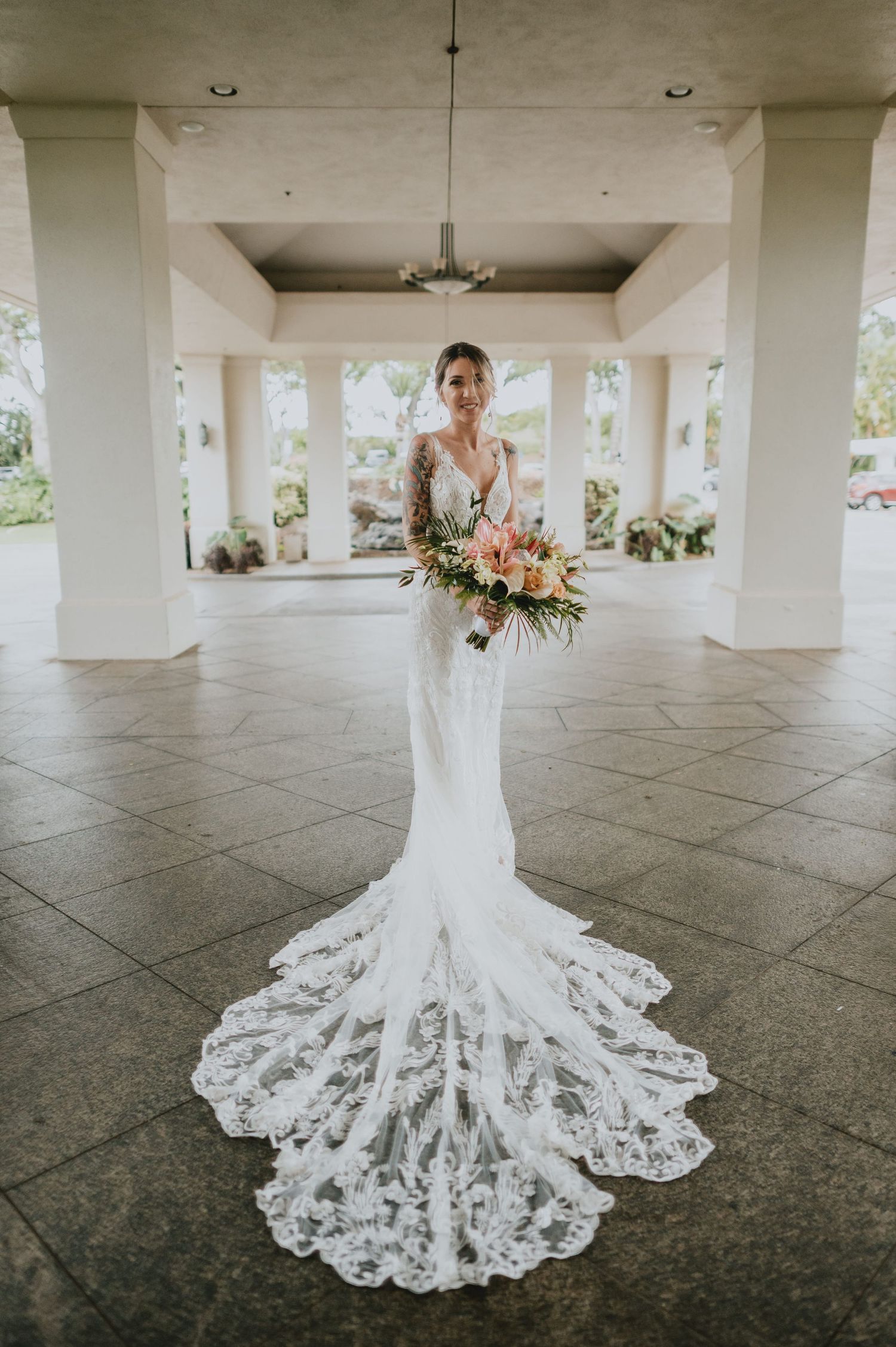Bride in elegant lace mermaid wedding gown with long train holds tropical bouquet under open-air venue columns.