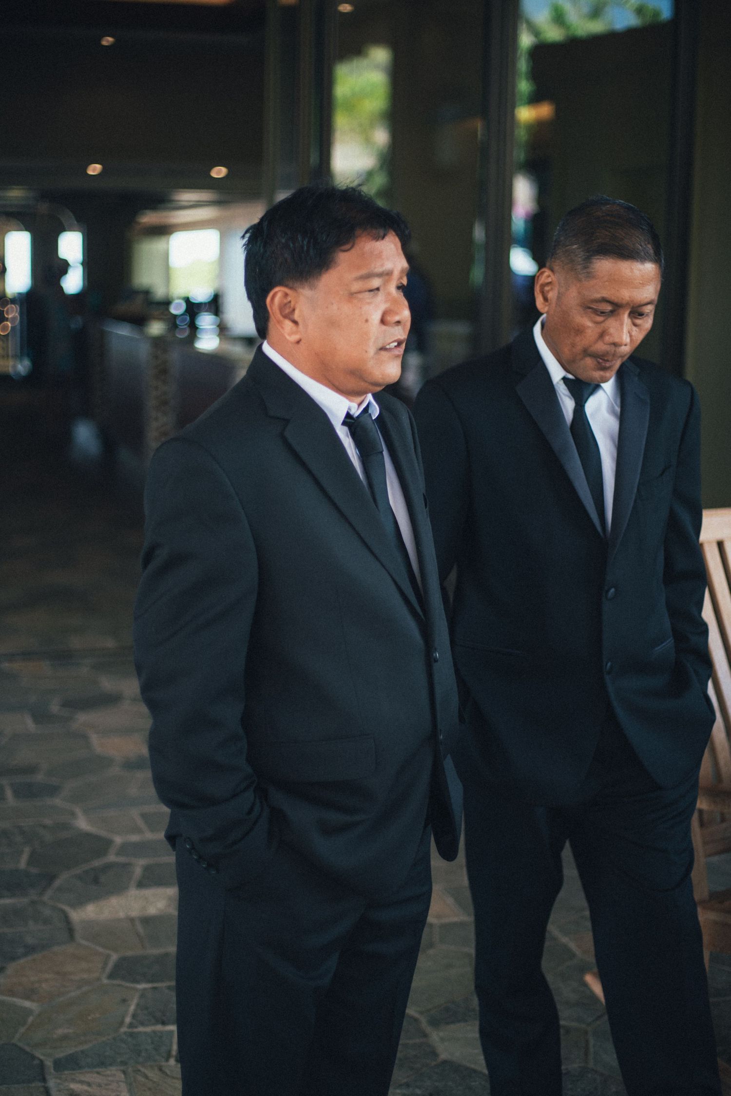 Two men in black suits and ties stand together in a formal setting with stone flooring.