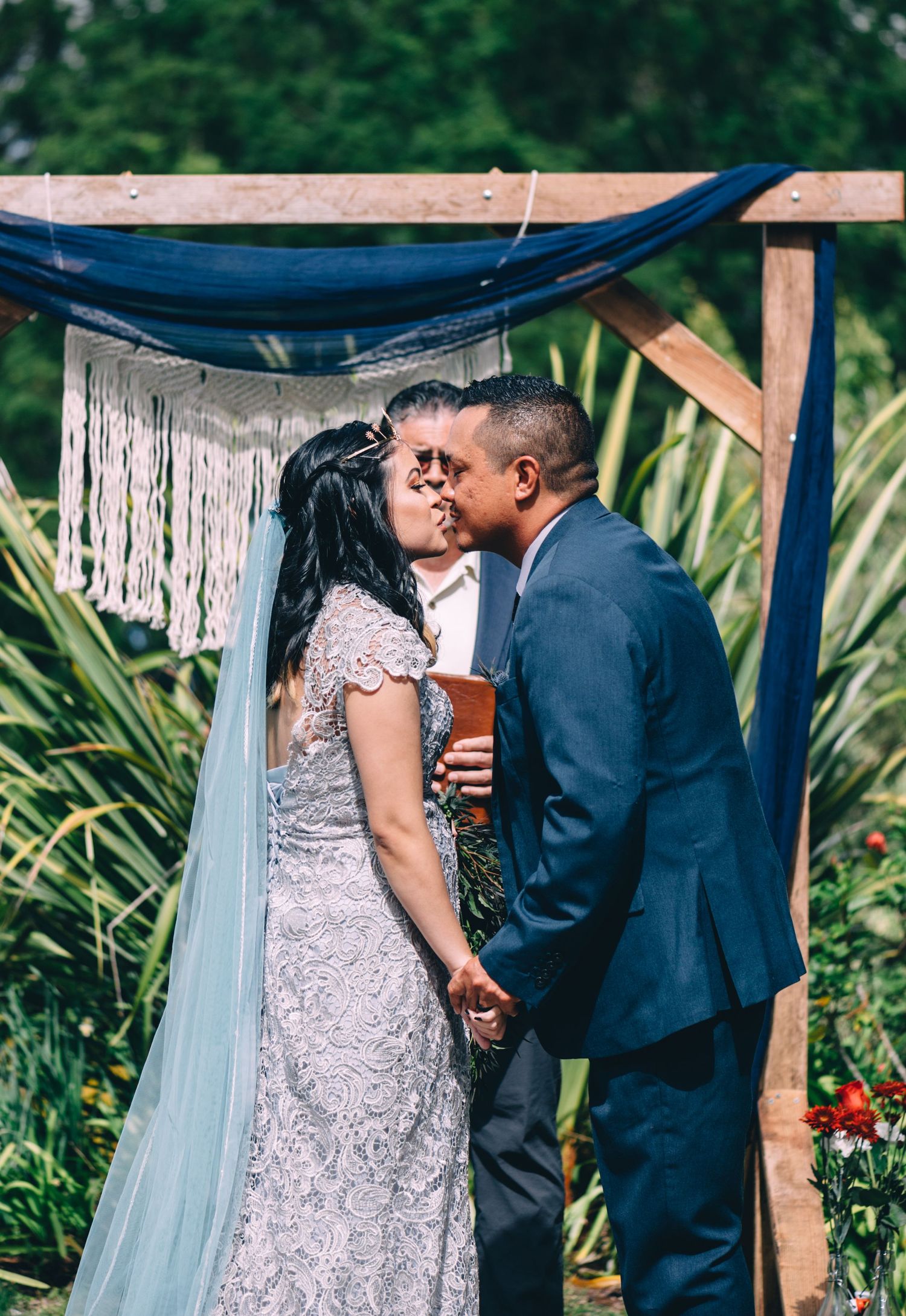 An intimate outdoor wedding ceremony moment captured under a rustic wooden pergola with blue fabric.