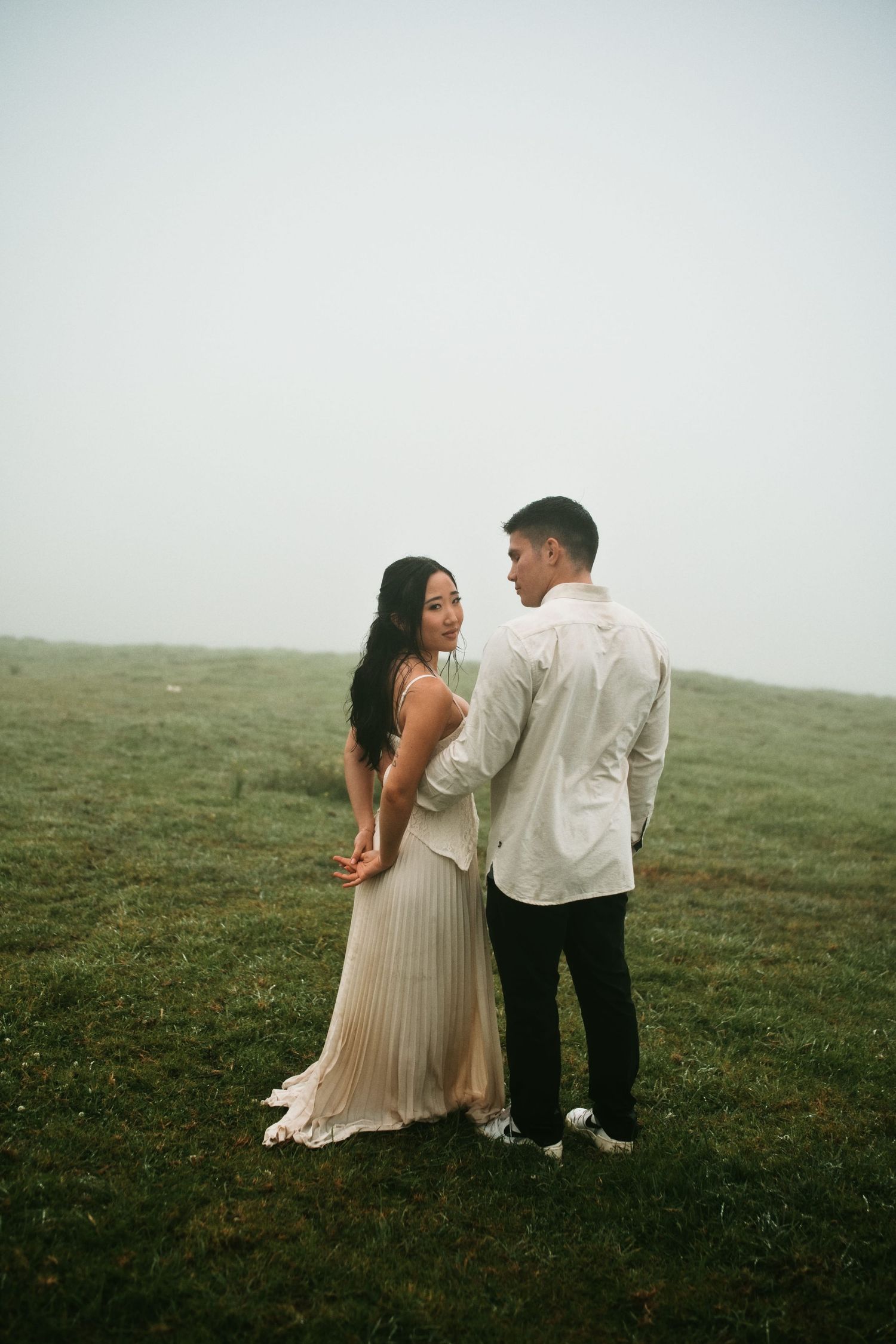 Couple holding hands in a misty green field with woman in elegant cream dress.