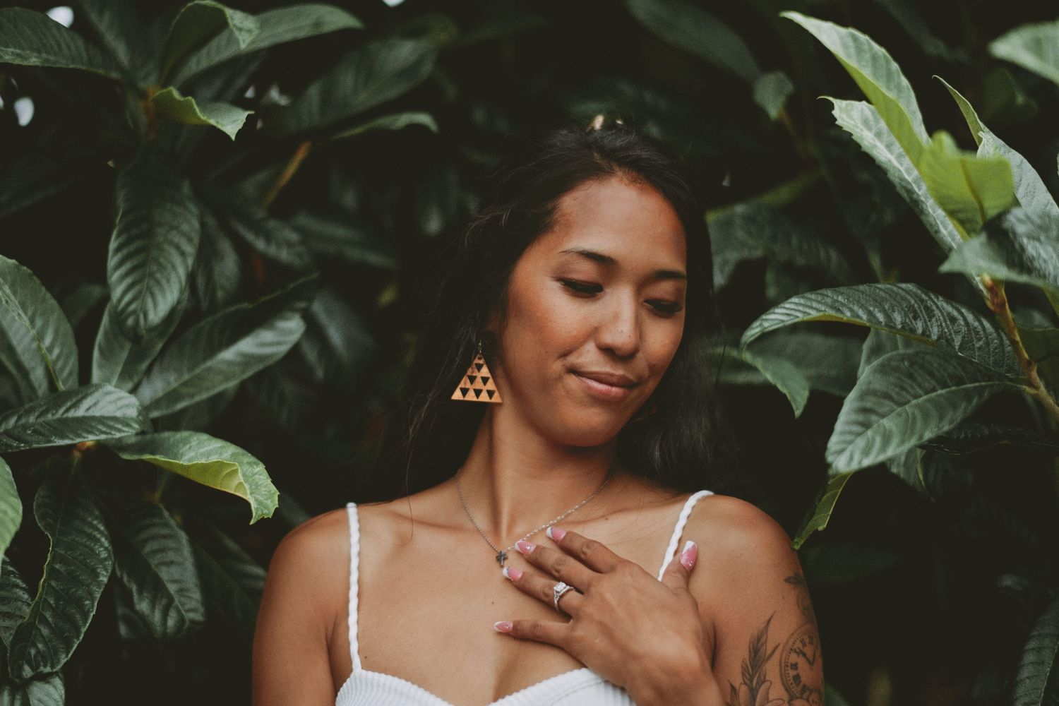 Woman in white dress with tattoo posing serenely among large tropical green leaves.