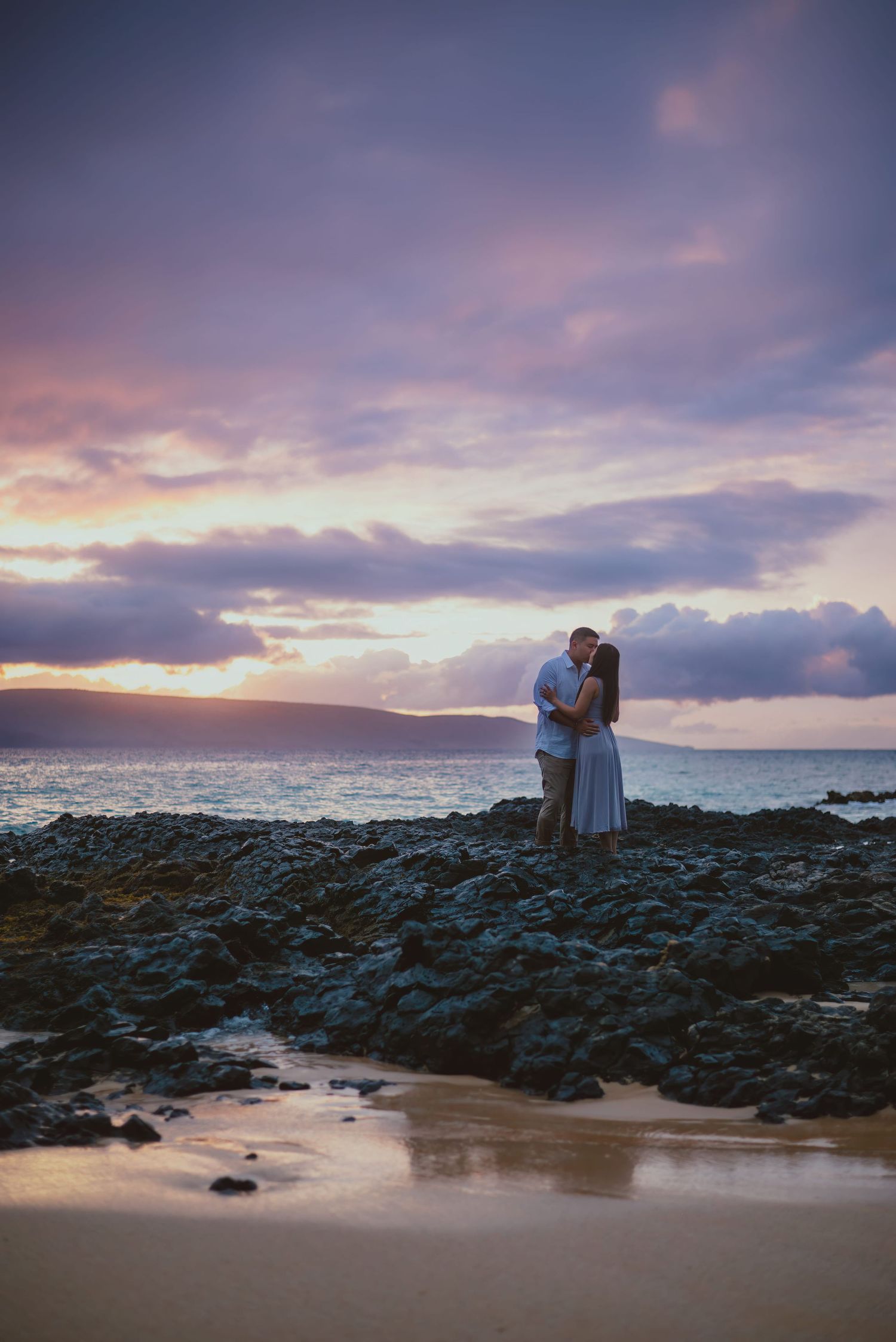 Couple kissing on rocky shore beneath a dramatic purple sunset sky in Maui Hawaii.