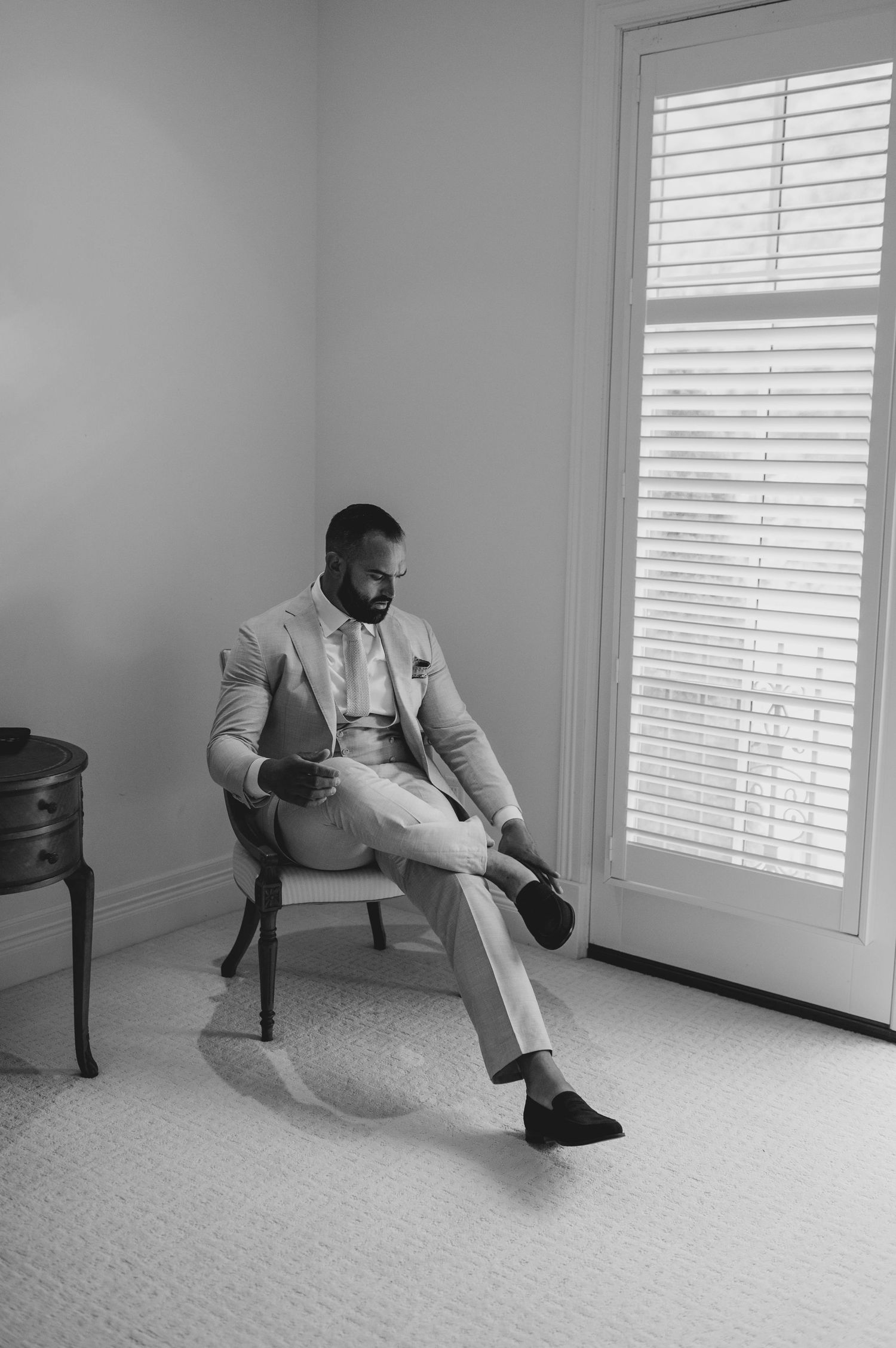 Dapper groom in light suit sitting in chair by window in black and white portrait.