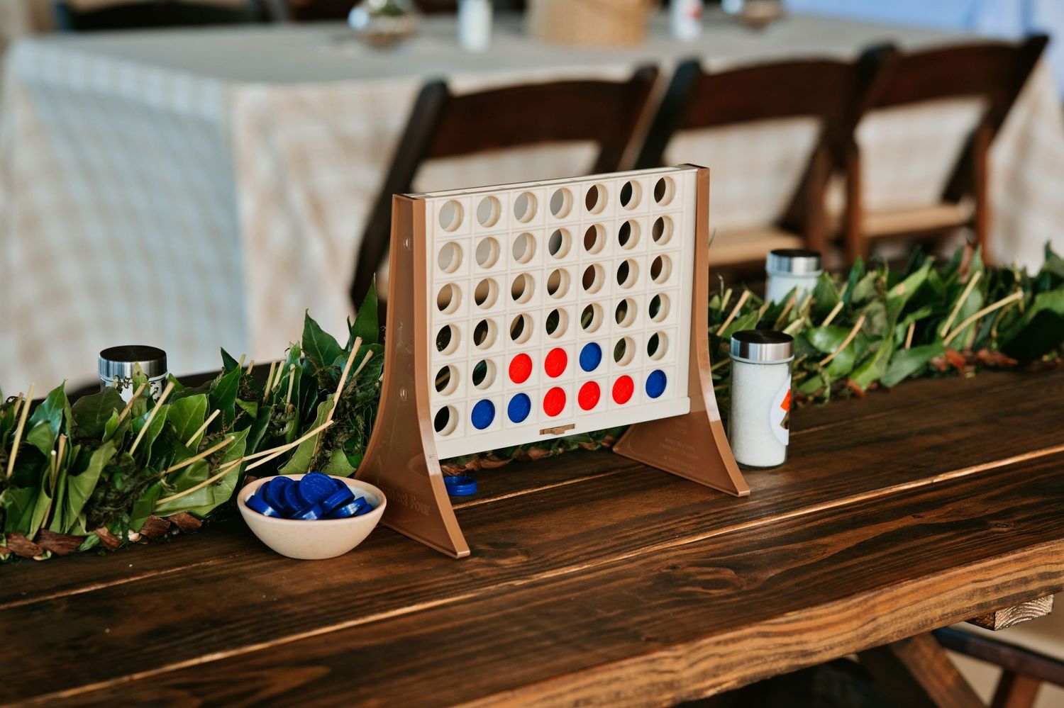 Giant Connect Four game on wooden table with garland, blue pieces bowl, at rustic wedding reception venue.