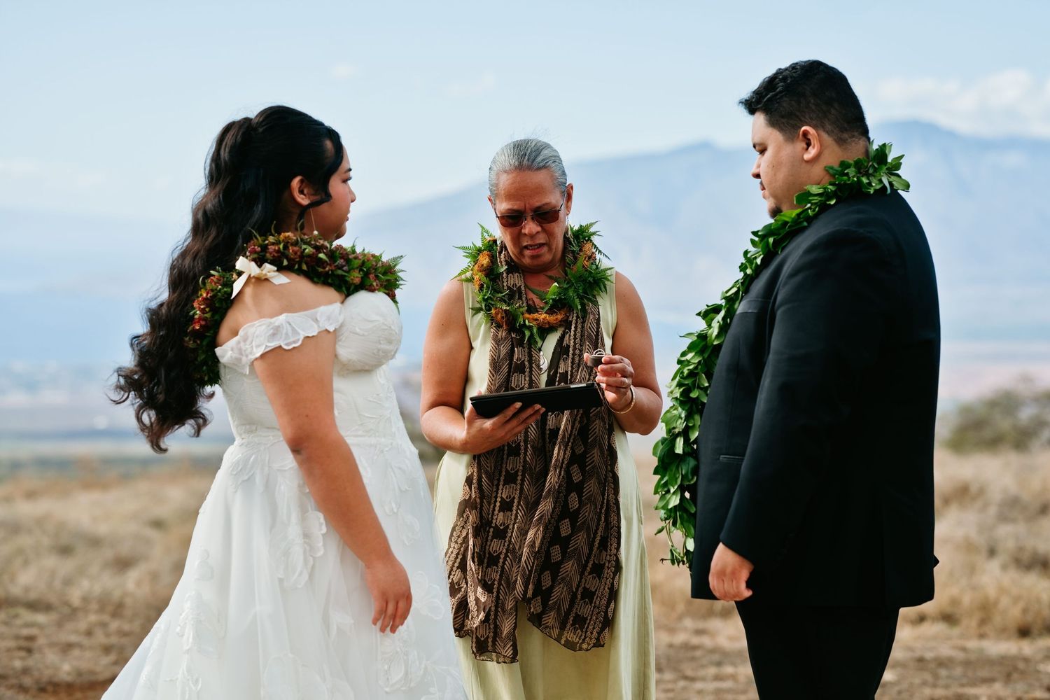 Couple in Hawaiian lei exchanging vows with officiant outdoors in Maui landscape during traditional wedding ceremony.