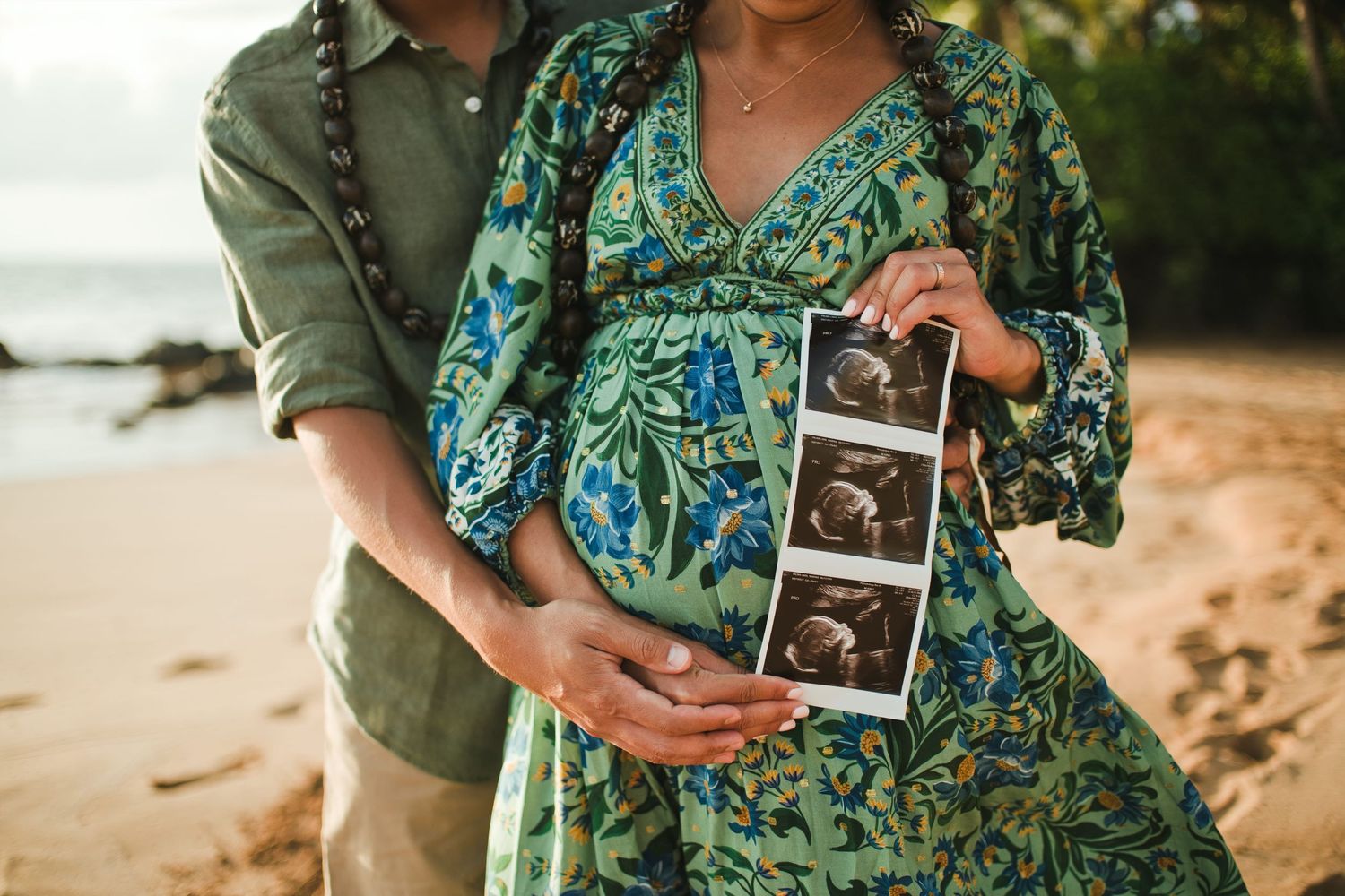 Pregnant couple on beach holding ultrasound photos, woman in floral green dress with partner embracing her baby bump.