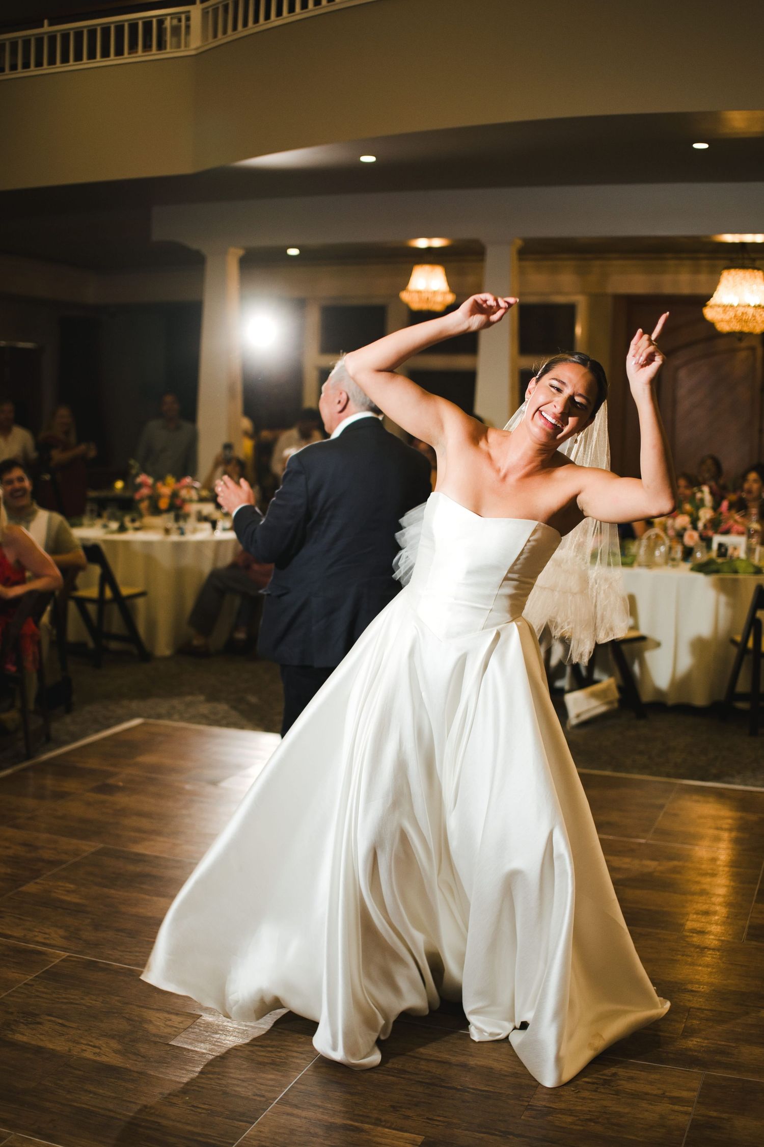 Joyful bride in white strapless gown dancing energetically at wedding reception with guests seated at tables in background.