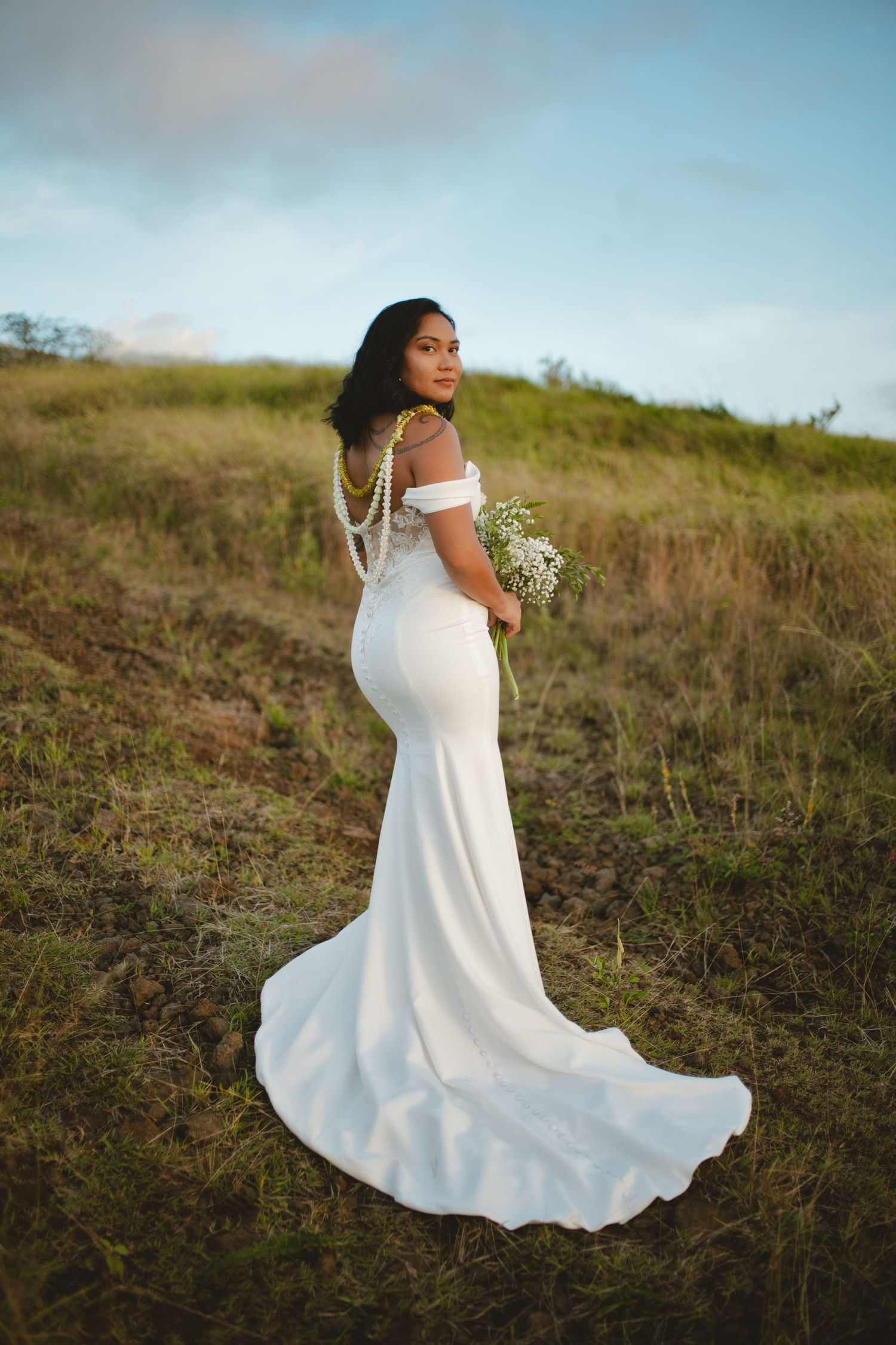 Bride in elegant off-shoulder white mermaid gown holds bouquet on lush green hillside.