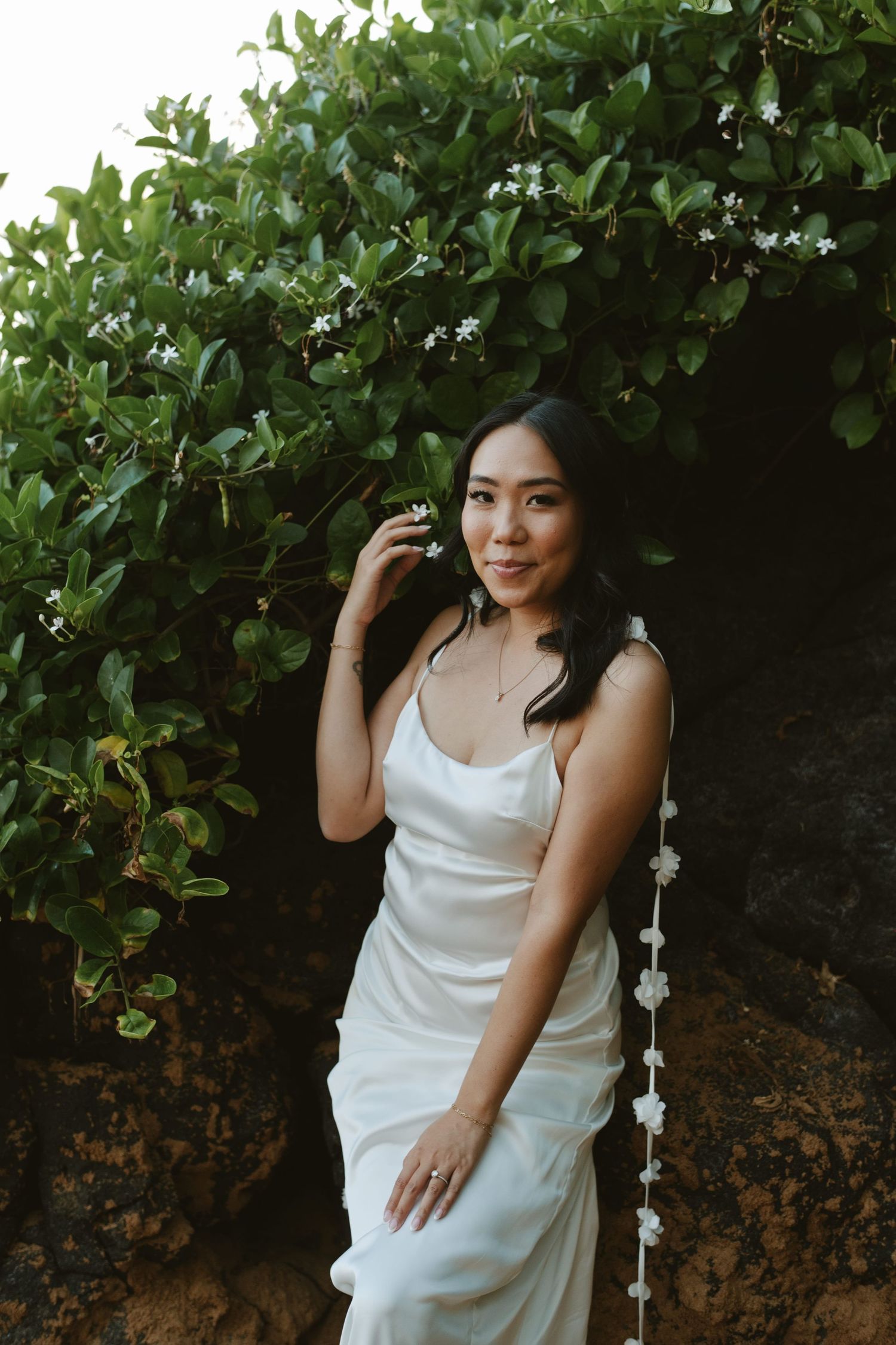 Bride in white satin gown posing under flowering green archway with floral hair accessory.