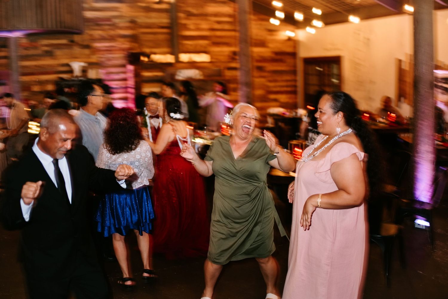 Guests dancing joyfully at a wedding reception in a rustic barn venue with warm lighting.