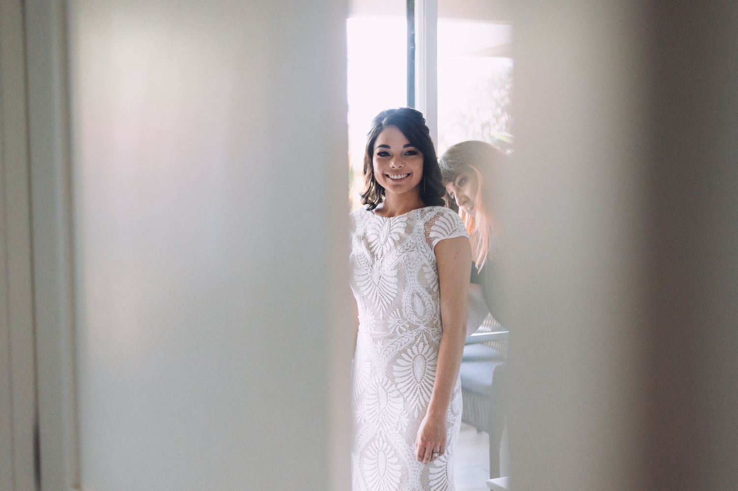 A bride in a lace wedding gown poses in front of a bright window.