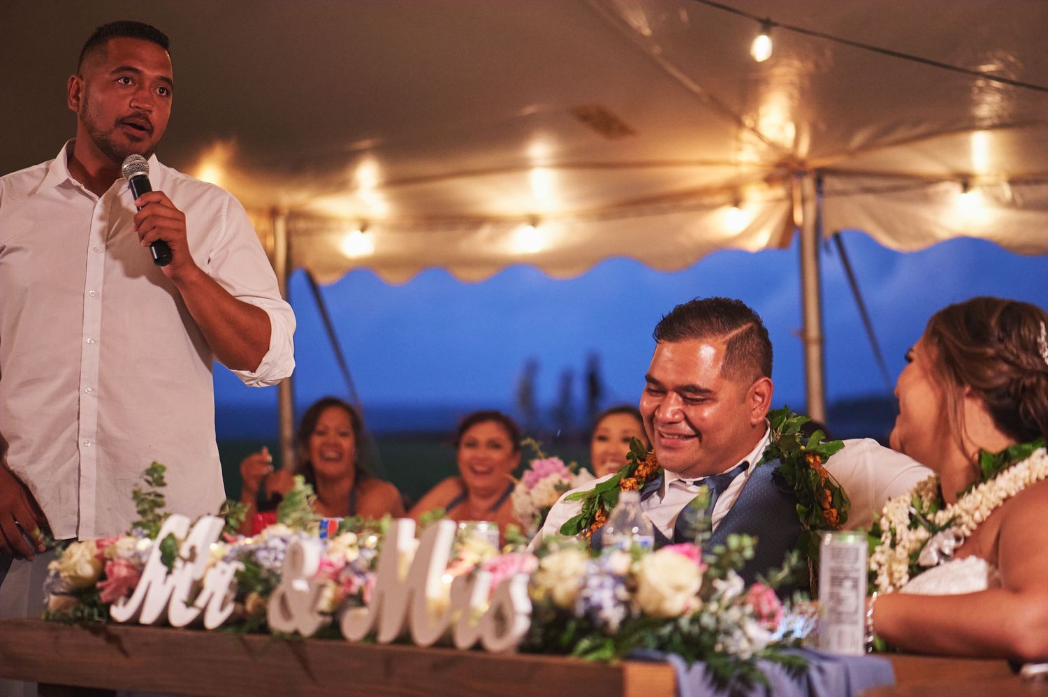 Wedding guests enjoy speeches and toasts during an outdoor evening reception under string lights.