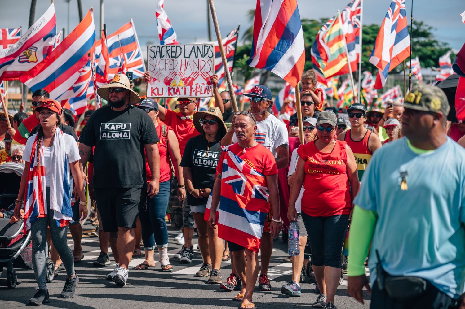Large group of demonstrators carrying Hawaiian flags march together during street protest.