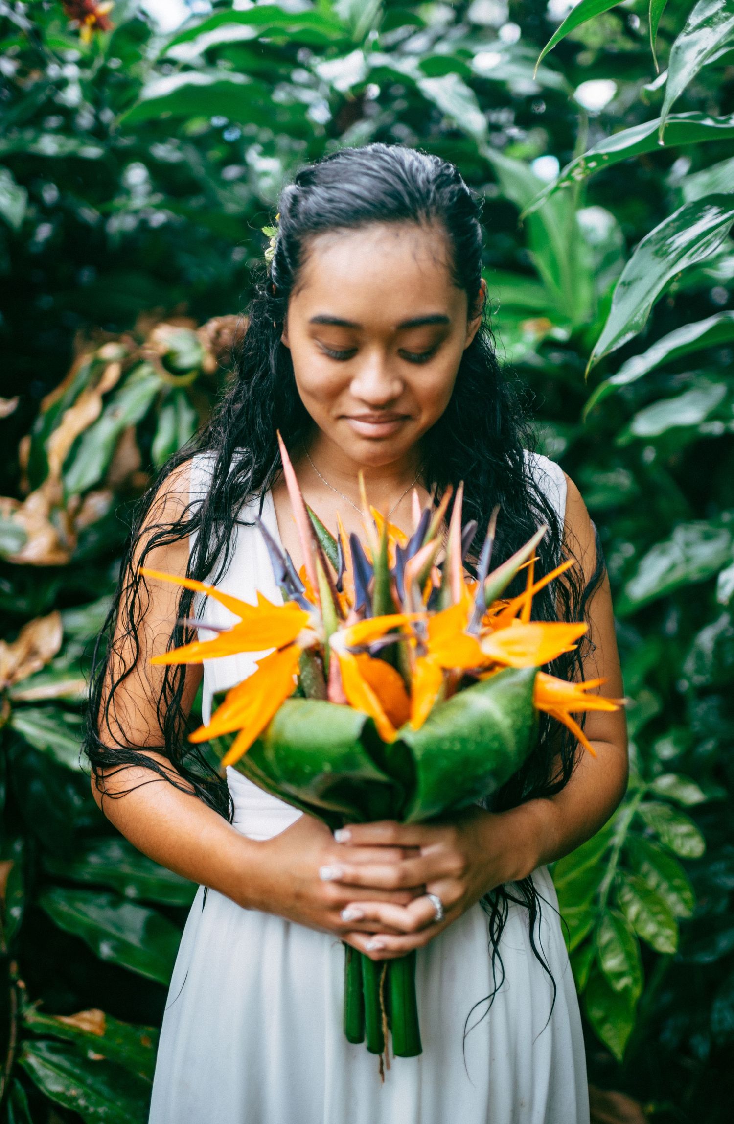 Young woman in white dress holding vibrant bird of paradise bouquet, smiling softly among lush tropical greenery.