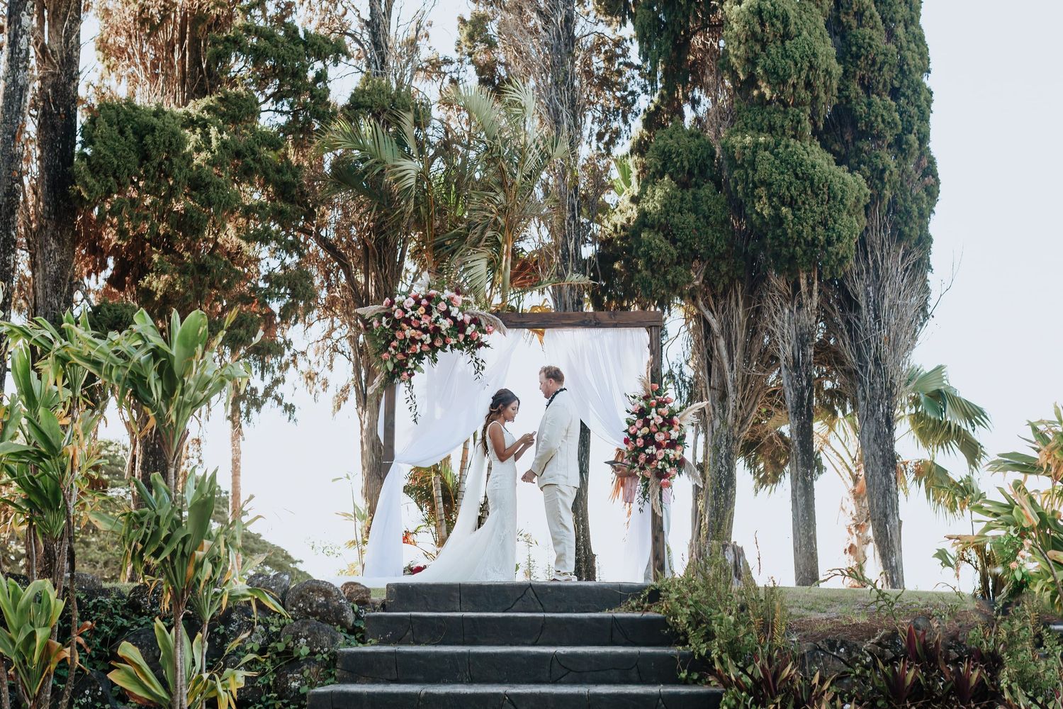 Couple exchanging vows at an outdoor wedding ceremony framed by lush tropical trees and floral arch.