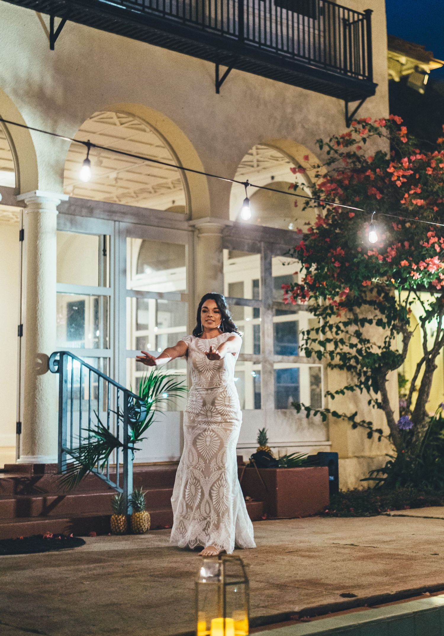 Bride in lace mermaid wedding gown posing outdoors at night in front of elegant building with string lights.