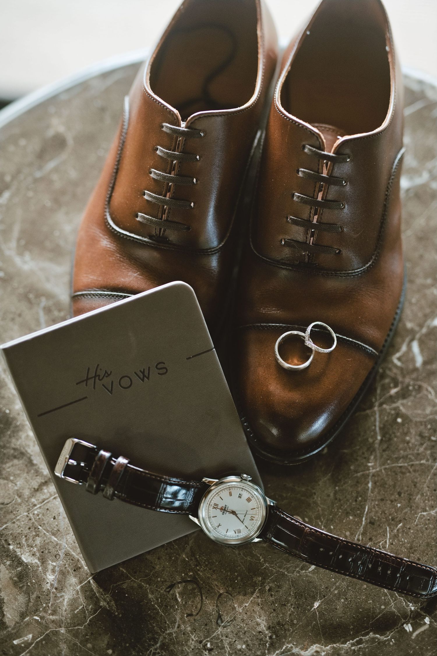 Brown leather oxford shoes, wedding rings, vows book, and classic wristwatch arranged on marble surface for groom wedding day details.