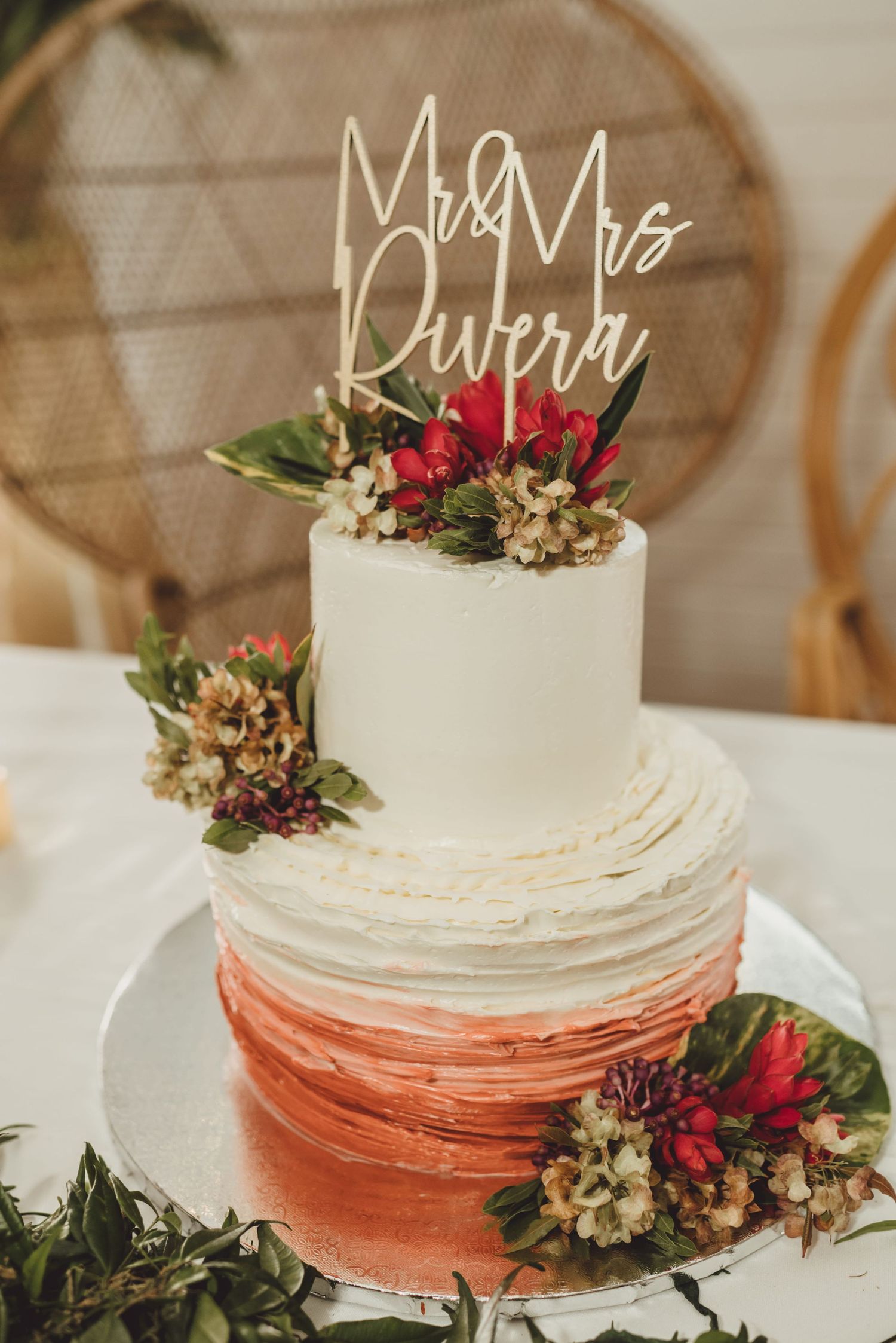 Two-tier white and coral ombre wedding cake with Mr & Mrs Rivera topper and tropical floral decorations.